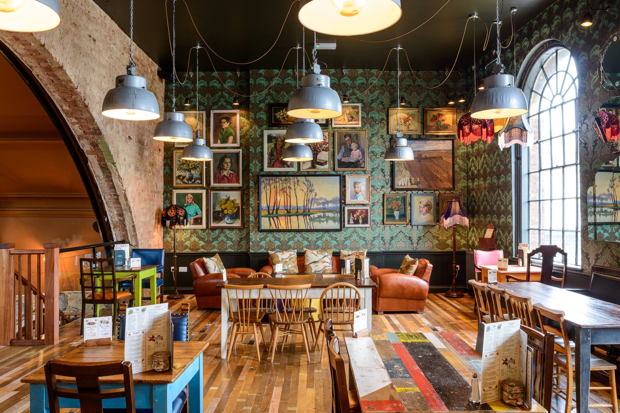 Cozy café interior with eclectic vintage decor, wooden furniture, Kino-inspired touches, colorful patterned walls adorned with framed artworks, pendant lights hanging from the ceiling, and a large arched window letting in natural light.