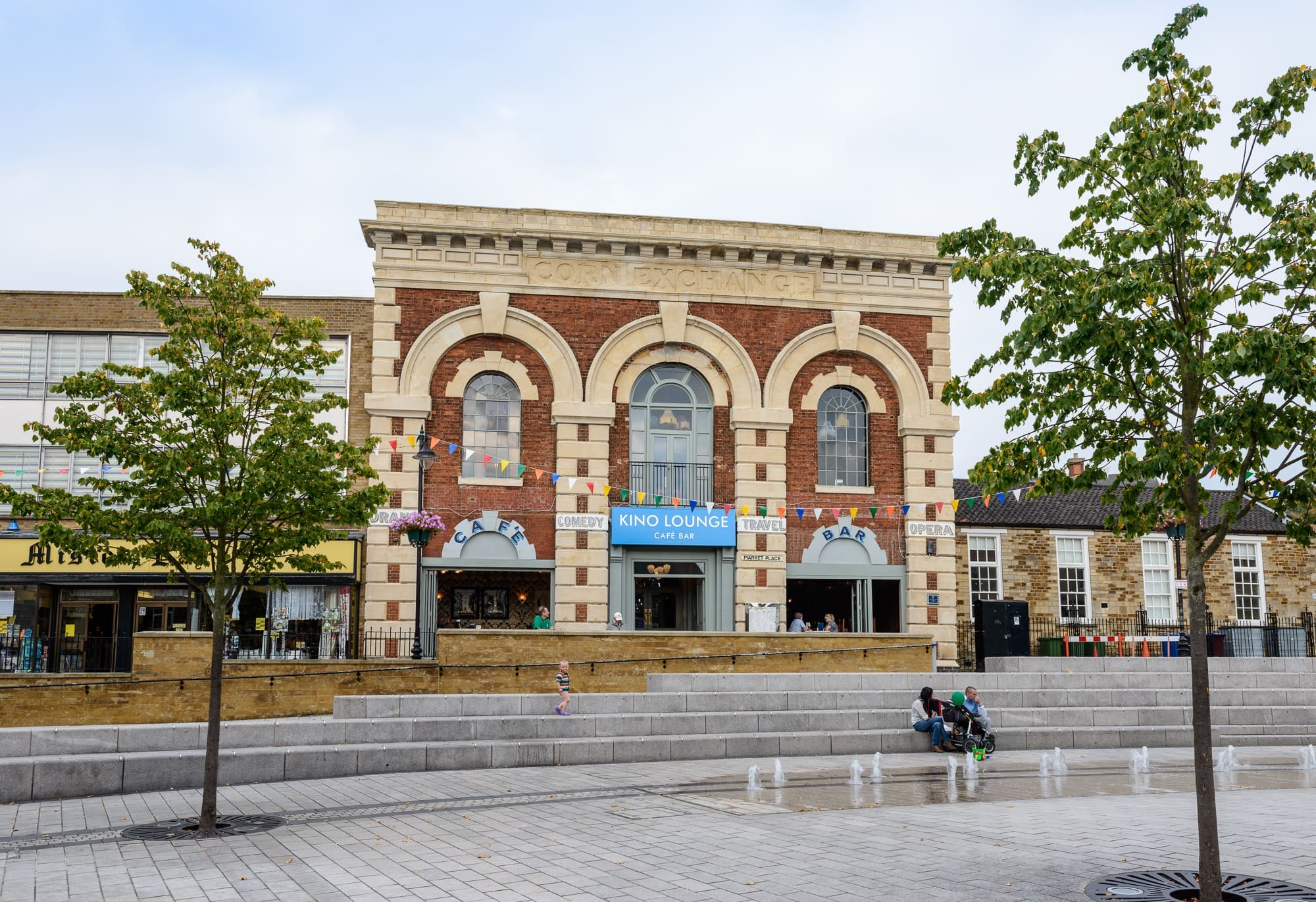 A historic brick building with arched windows houses the Kino Lounge café. Bunting hangs above the entrance, and people relax near water fountains in a modern paved plaza with young trees, creating a vibrant spot for Kino visitors.