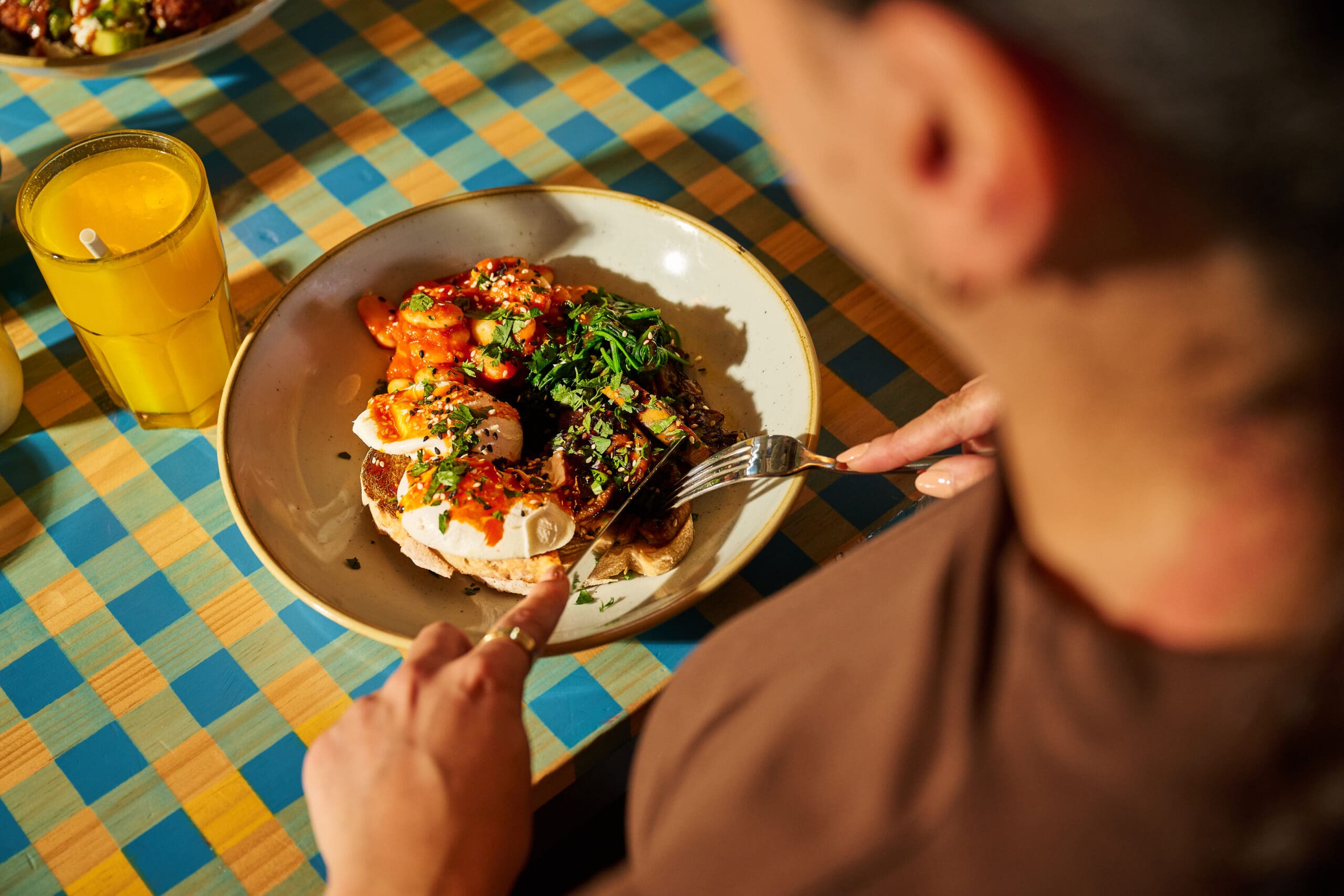 A person holding a fork and knife prepares to eat a colorful dish with greens, sauce, and bread, accompanied by a glass of orange juice, on a checkered tablecloth.