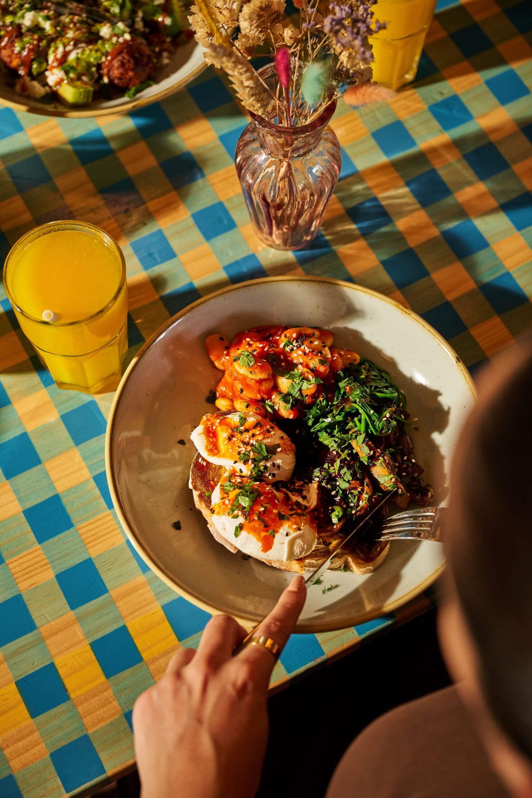 A person holding a fork sits at a table with a plate of food featuring grilled chicken topped with sauce and herbs, sautéed greens, and grains. A glass of orange juice and a vase with dried flowers are also on the checkered tablecloth.