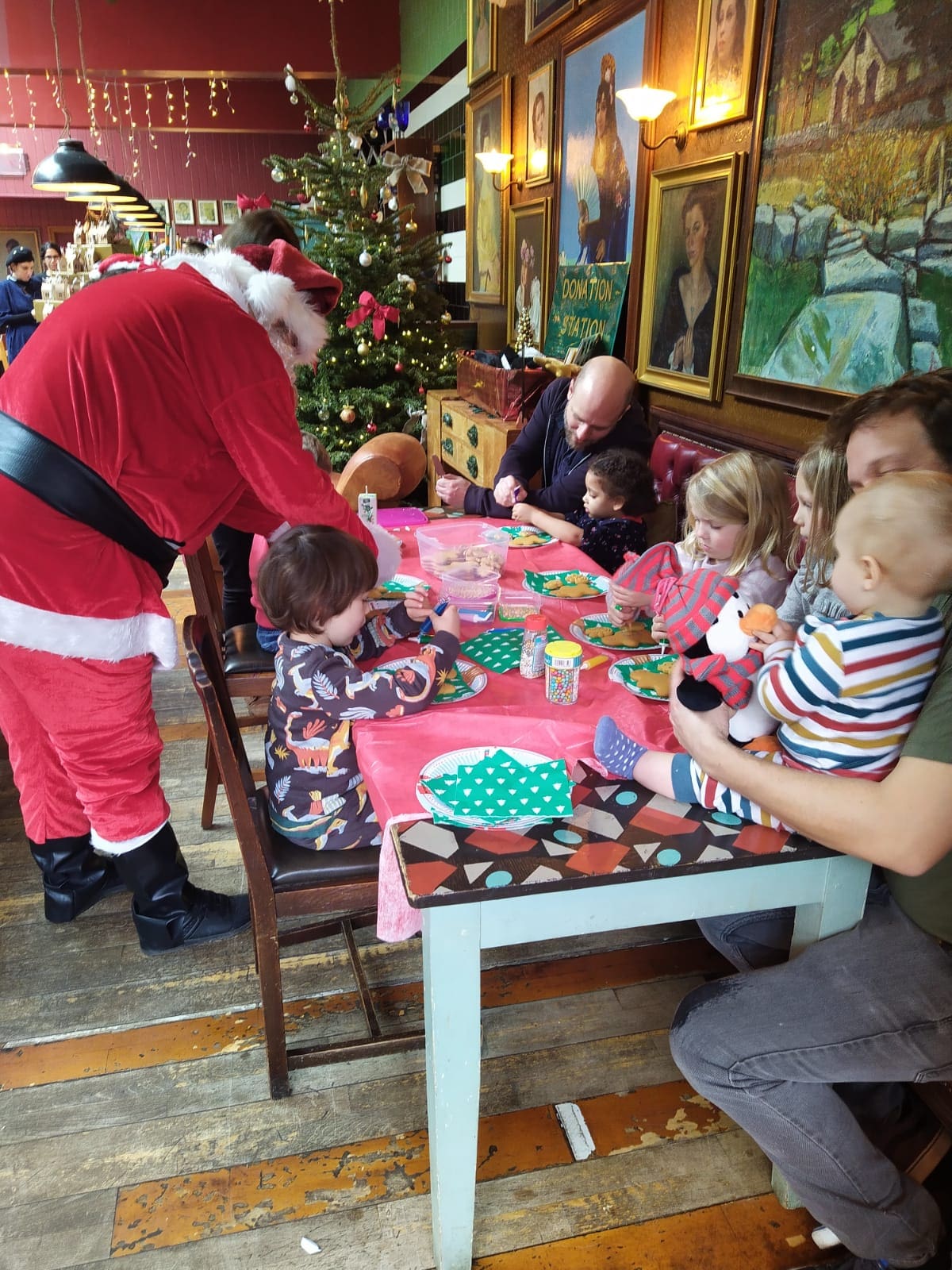Santa Claus interacts with children seated around a festive table, decorated with Christmas-themed items. A Christmas tree and holiday decorations are visible in the background, with adults and children enjoying the event.