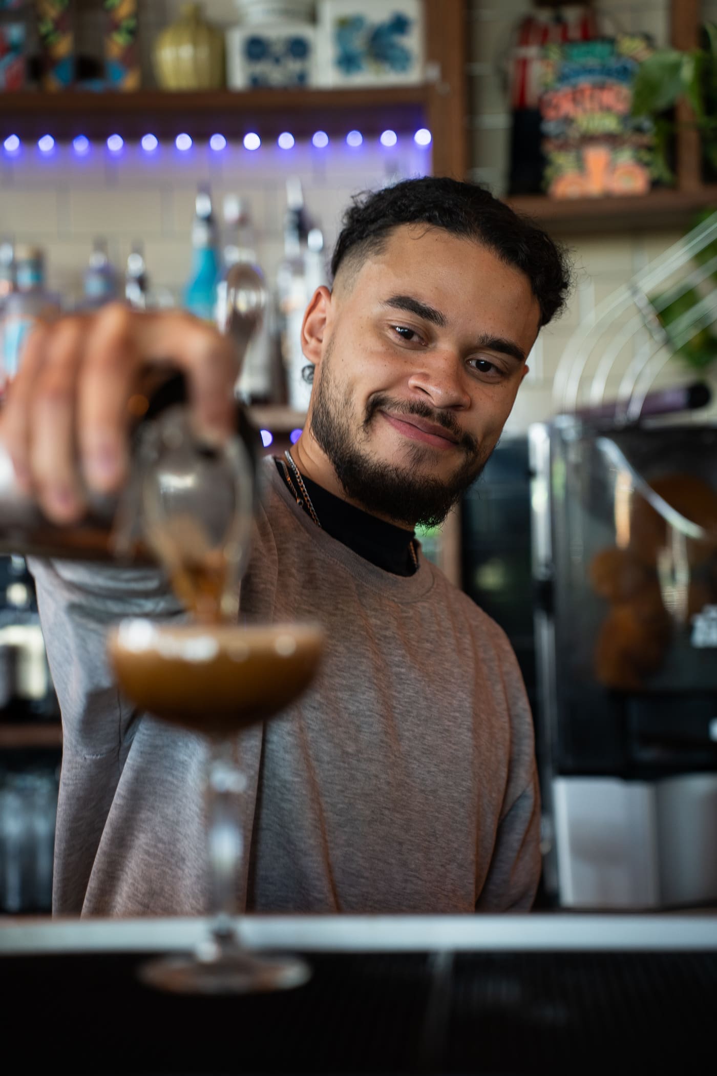 A smiling bartender with a beard pours a brown cocktail into a glass at a bar, highlighting the vibrant atmosphere and exciting careers available in the hospitality industry, with bottles and colorful decor visible in the background.