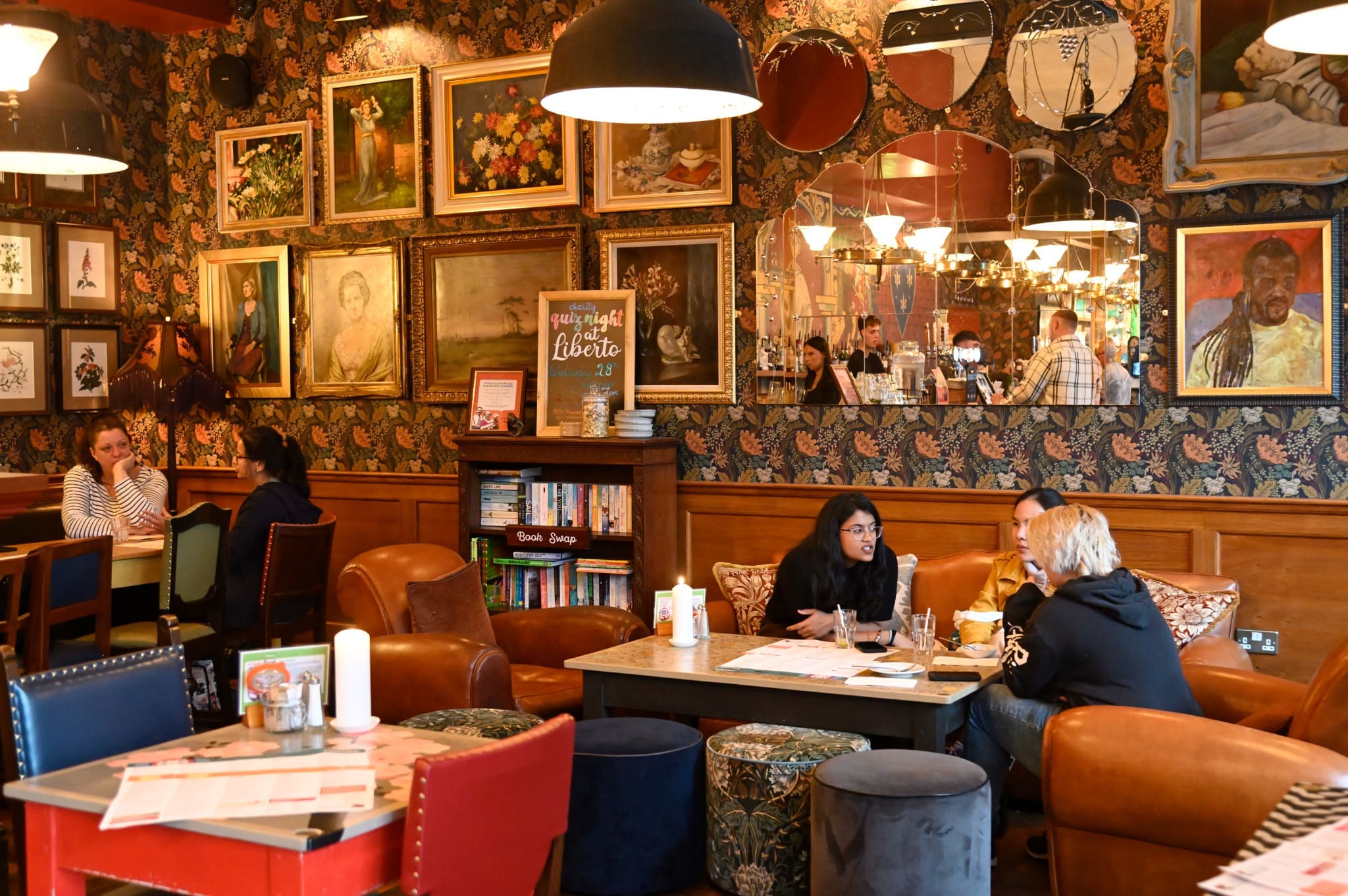 A cozy café with people sitting at tables, chatting and eating. The walls are decorated with Liberto paintings and mirrors, and bookshelves filled with books create a warm, inviting atmosphere.