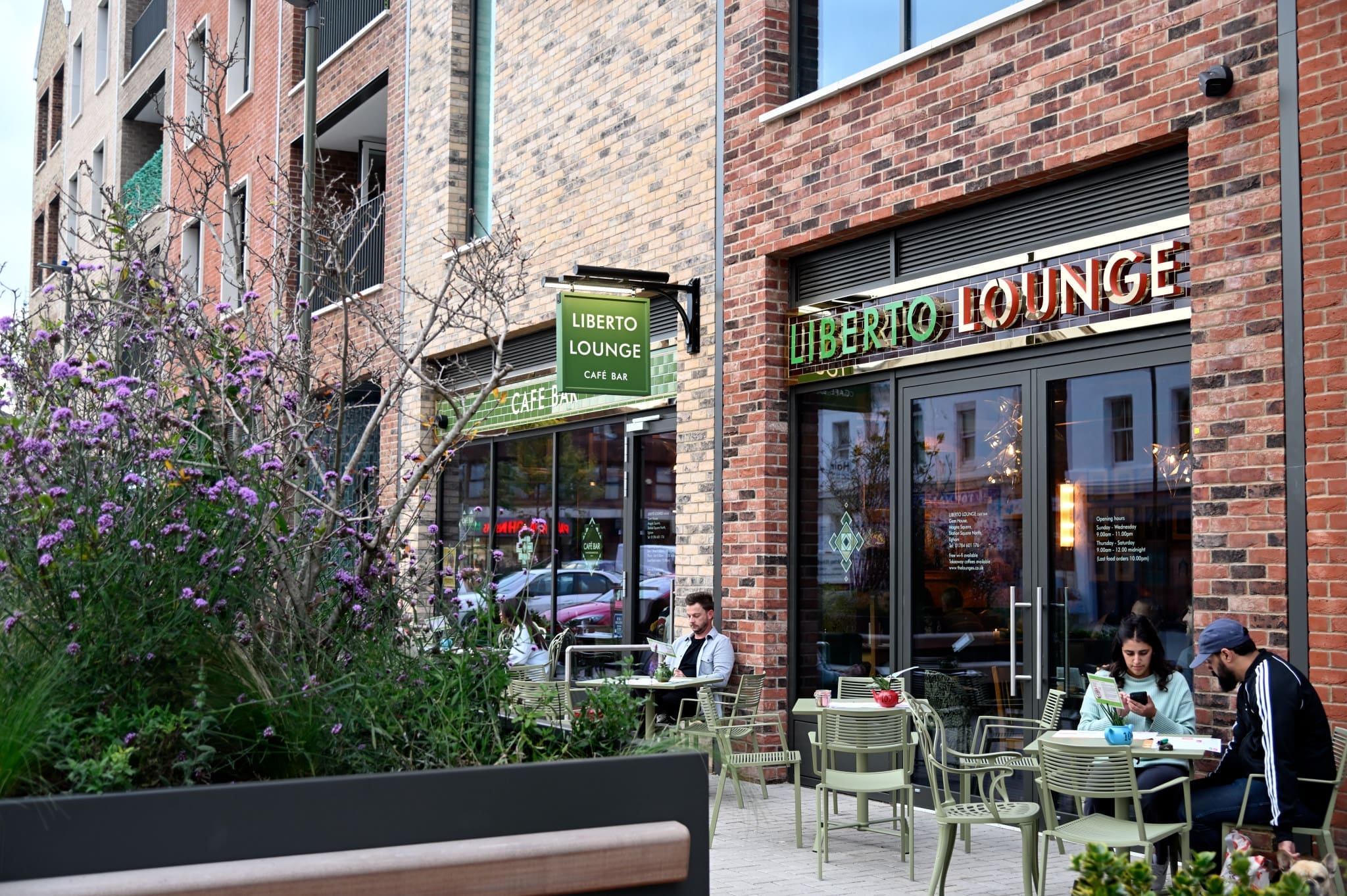 People sit at outdoor tables in front of Liberto Lounge café, a modern brick building with large windows and green signage. Potted plants and purple flowers decorate the area, while the inviting Liberto atmosphere draws in passersby.