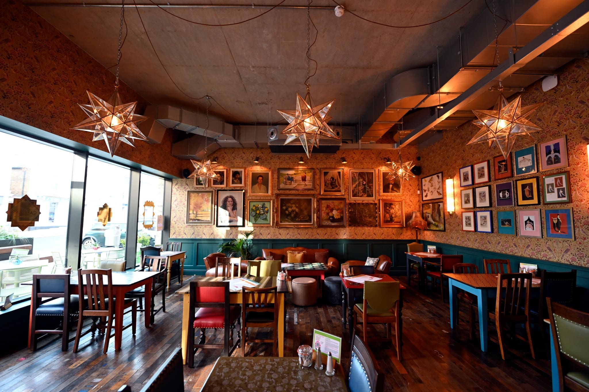 A cozy, eclectic café by Liberto with colorful mismatched chairs and tables, star-shaped pendant lights, wooden floors, and art-filled walls featuring paintings and portraits, with large windows letting in natural light.