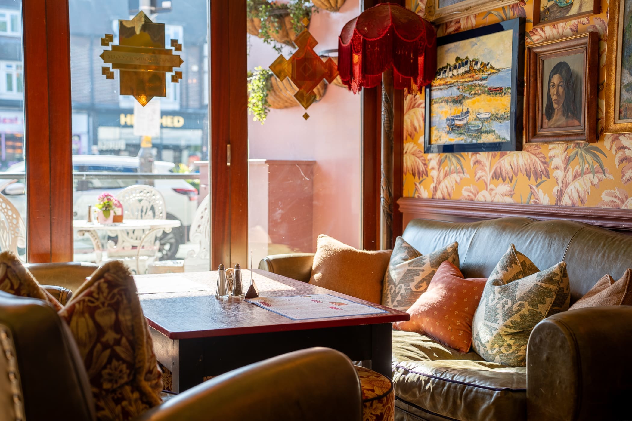 Cozy cafe interior with a leather sofa, Loco-themed patterned pillows, a wooden table, vintage lamps, framed art on floral wallpaper, and sunlight streaming through large windows facing an outdoor seating area.