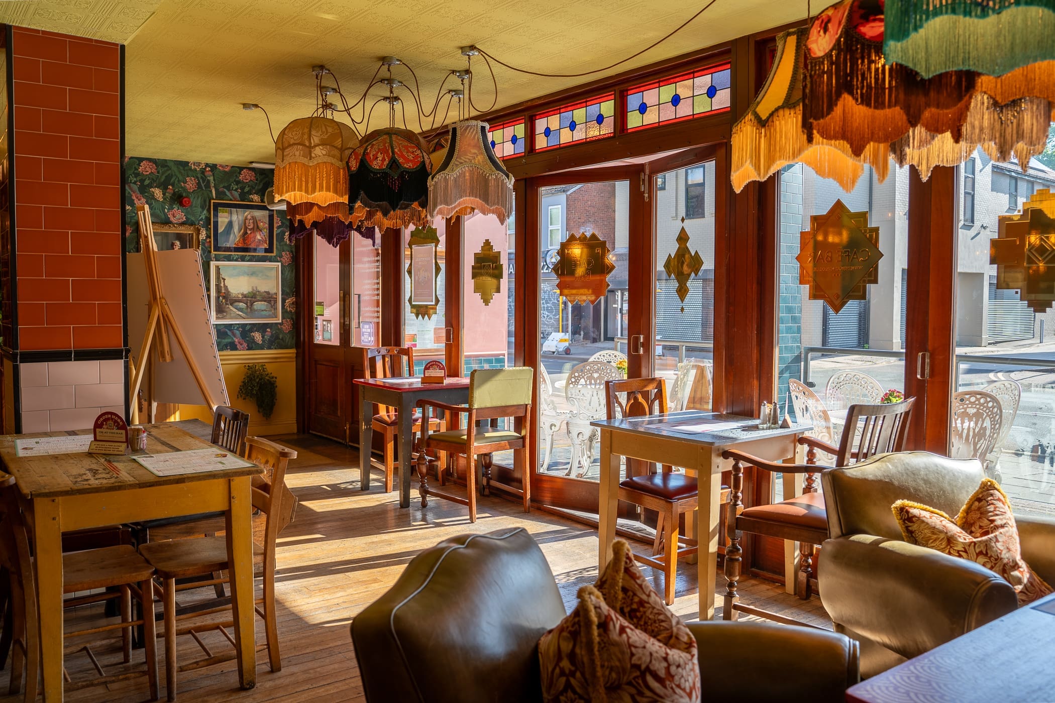 A cozy café interior with vintage decor and a touch of Loco flair, colorful stained glass windows, fringed lamps, assorted tables and chairs, soft cushions, and sunlight streaming through large windows onto the wooden floor.