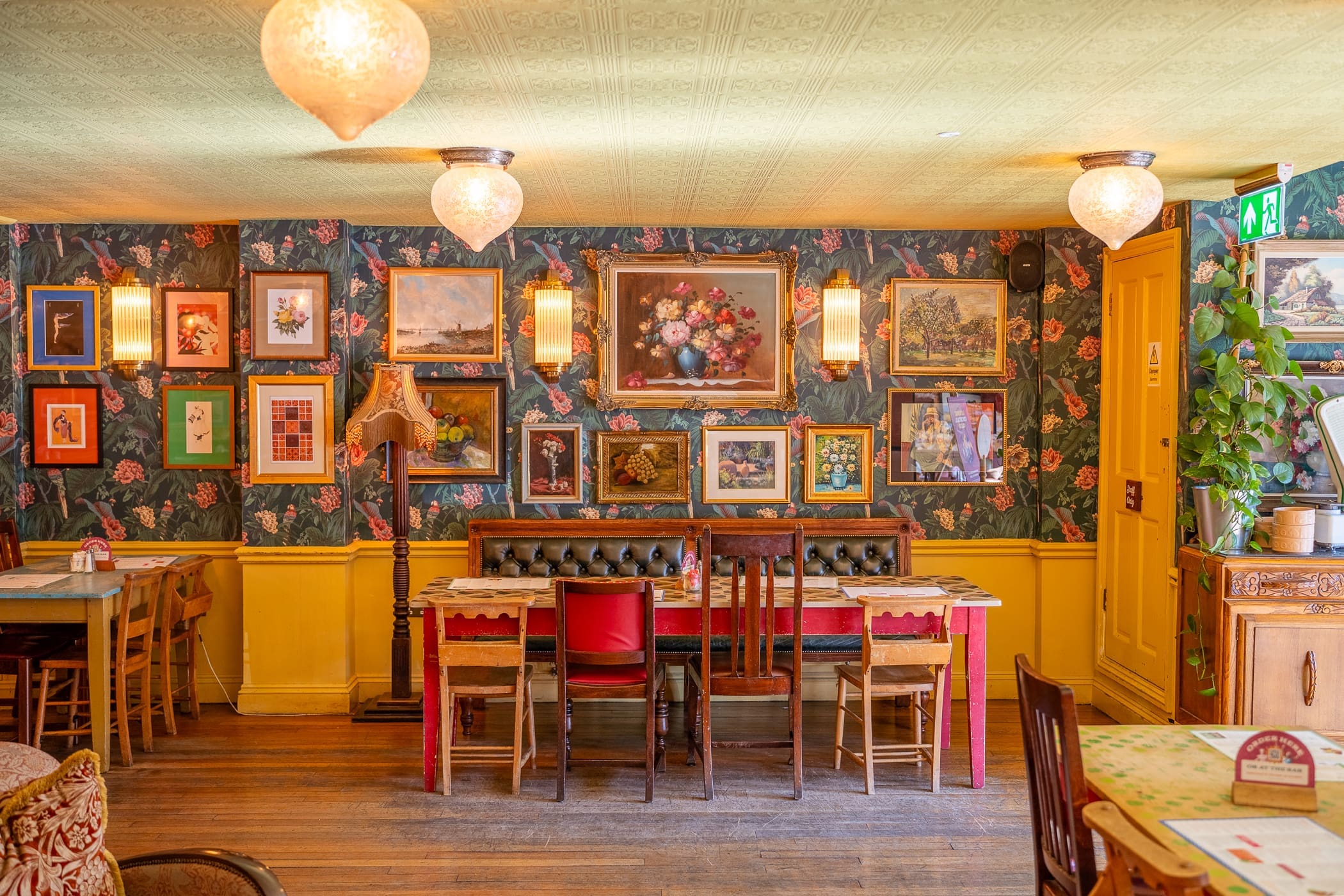 Cozy vintage-style café interior with floral wallpaper, assorted framed art, wooden tables and chairs, an antique lamp, and warm lighting—Loco charm meets inviting, eclectic atmosphere for a truly unique experience.