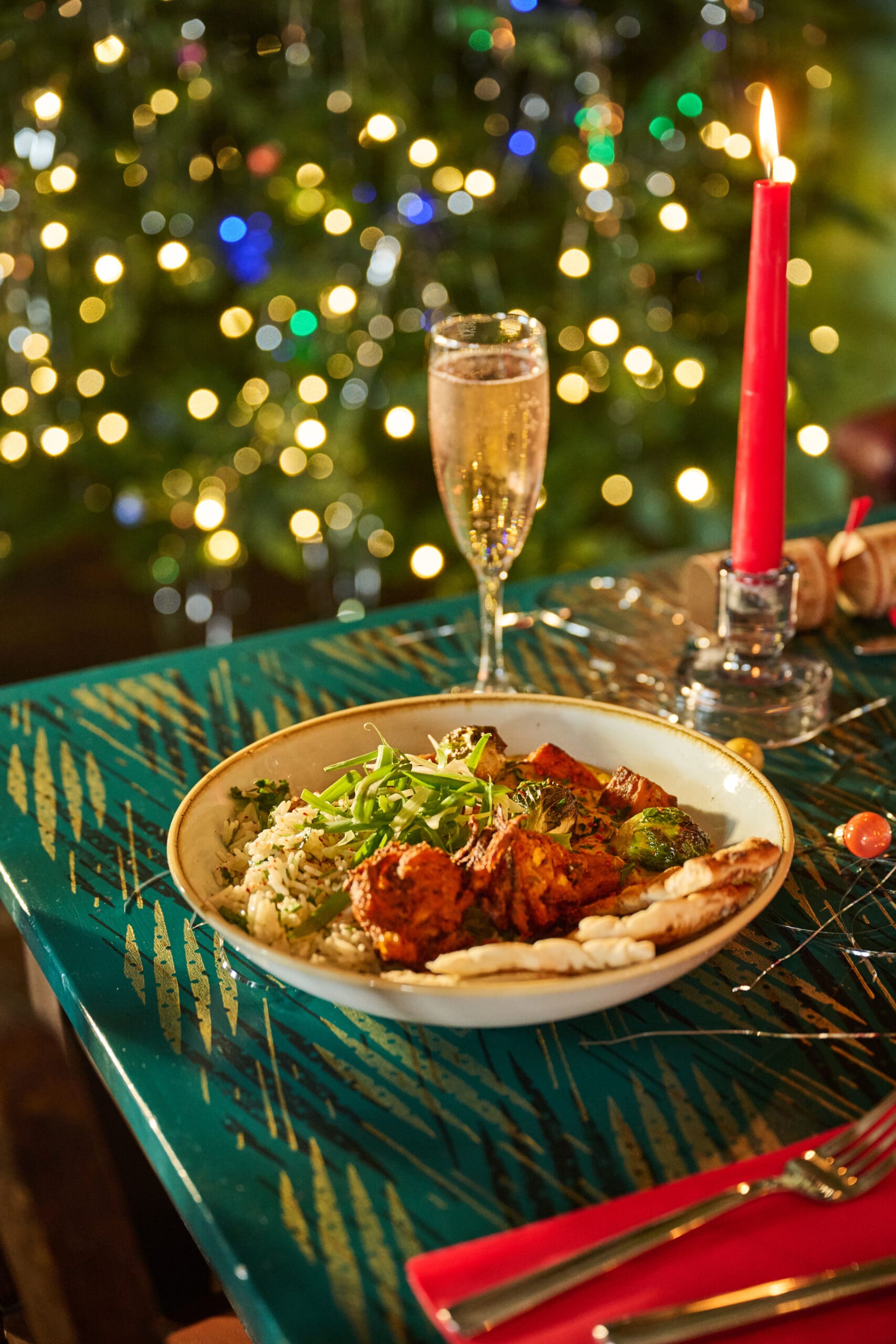 A festive table setting with a plate of food, a glass of champagne, and a lit red candle, in front of a decorated Christmas tree with colorful, blurred lights.