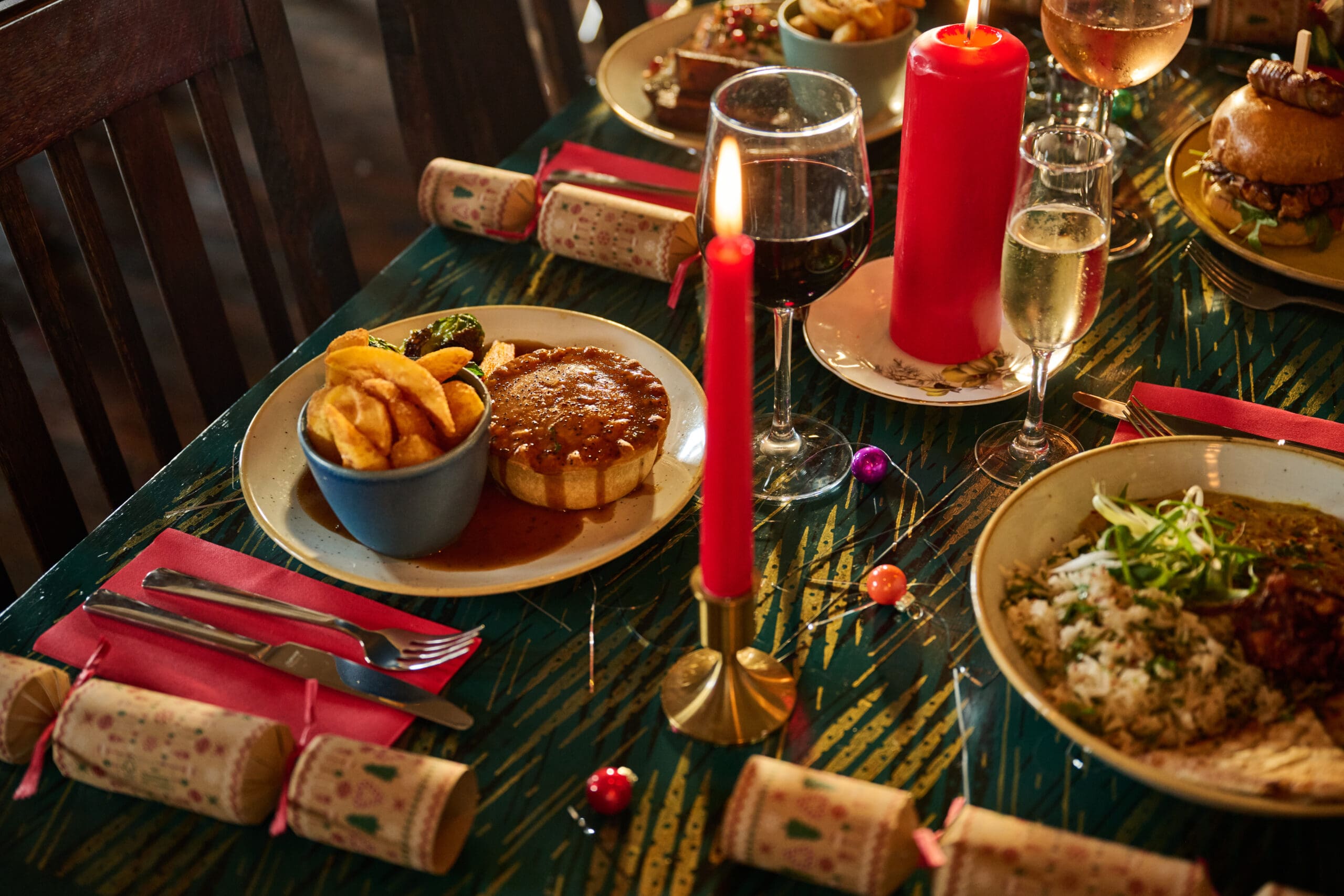 A festive dinner table set in a cozy lounge with plates of food, drinks, red candles, Christmas crackers, and decorations. Dishes include pie with gravy and chips, a rice dish, and a burger, creating a warm holiday atmosphere.