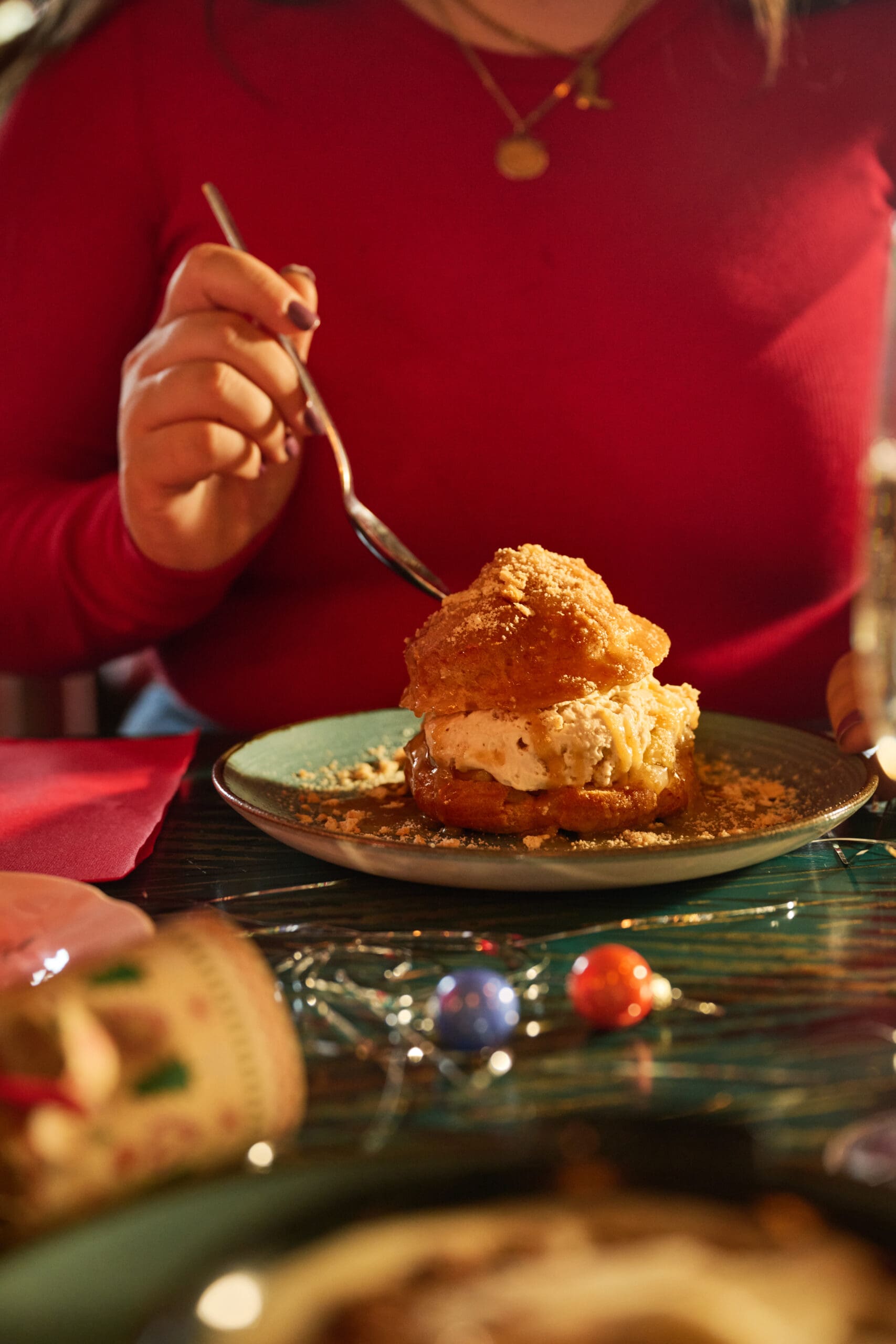 A person in a red top holds a spoon, about to eat a cream-filled pastry dusted with sugar on a plate. Holiday decorations and a festive ambiance are visible on the table.
