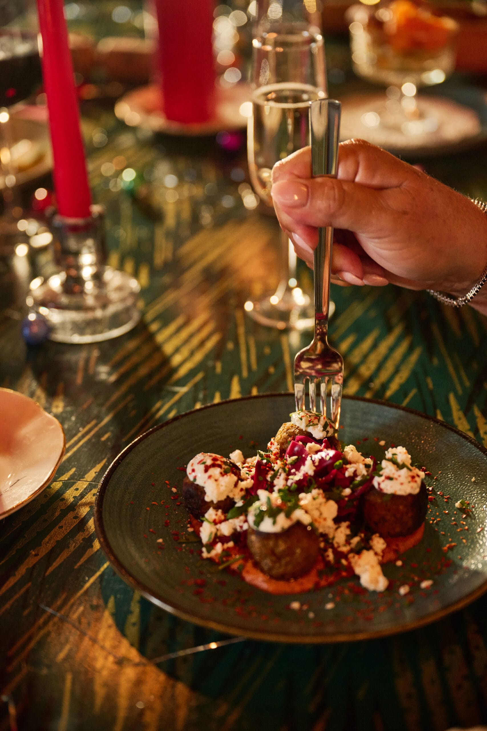 A hand holding a fork is about to eat an appetizer topped with crumbled cheese and herbs on a dark plate, set on a festive table with candles and glasses in the background.
