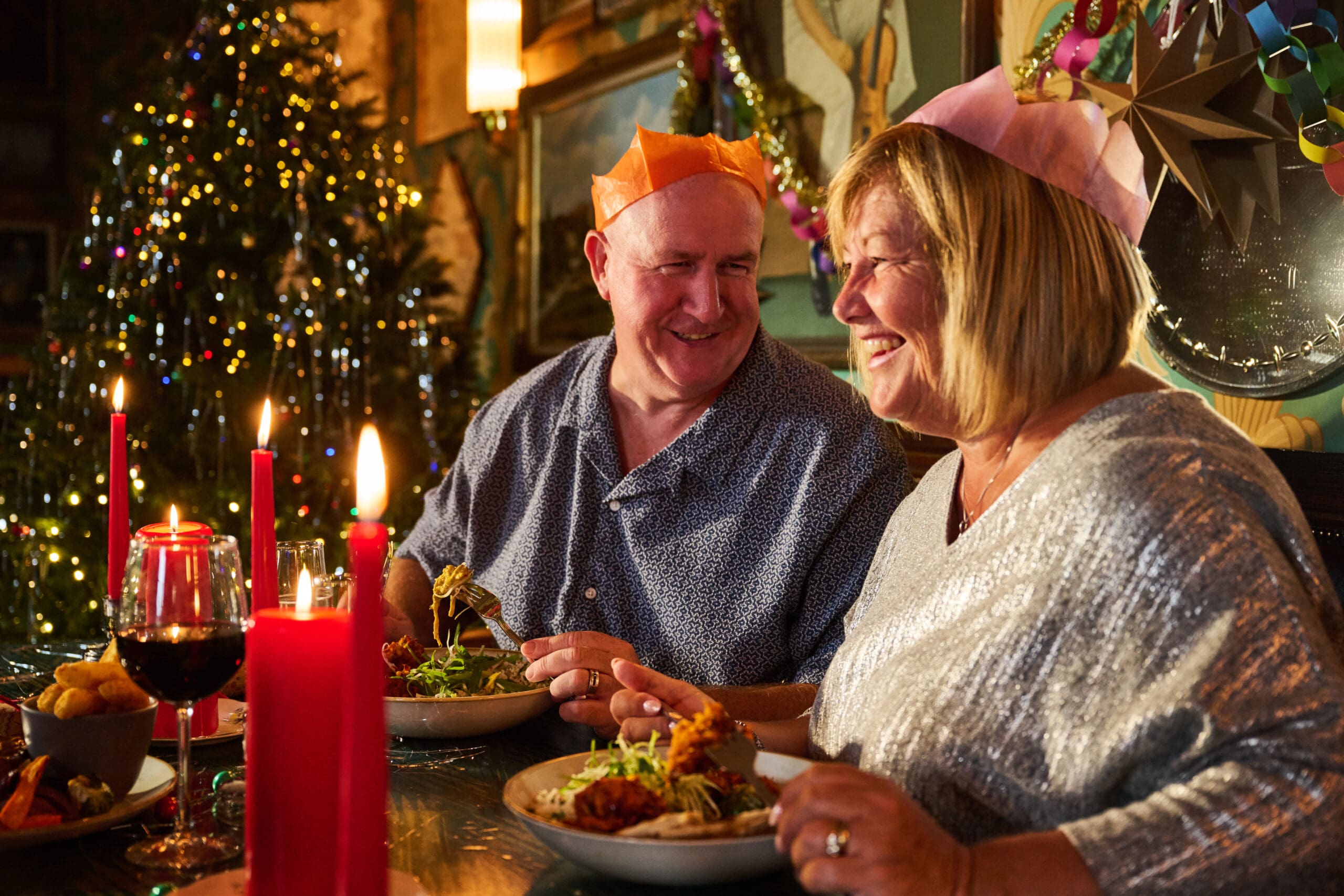 An older couple wearing colorful paper crowns enjoy a festive meal together at a decorated table, with a Christmas tree, lit candles, and food in the warm, cozy background.