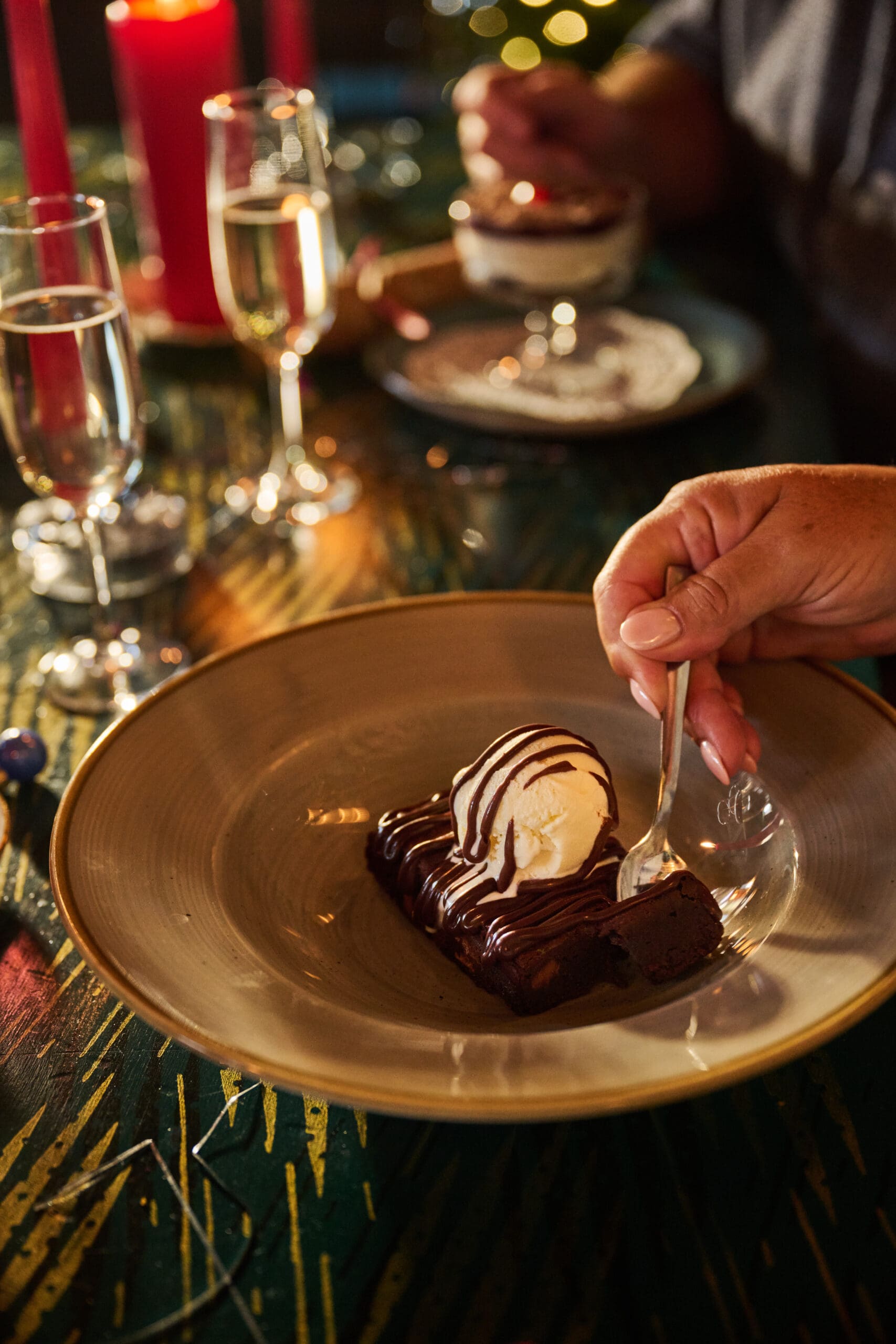 A hand holds a spoon about to eat a dessert of chocolate brownie topped with vanilla ice cream and chocolate sauce, served on a plate. Two glasses of sparkling wine and candles are visible in the dimly lit background.