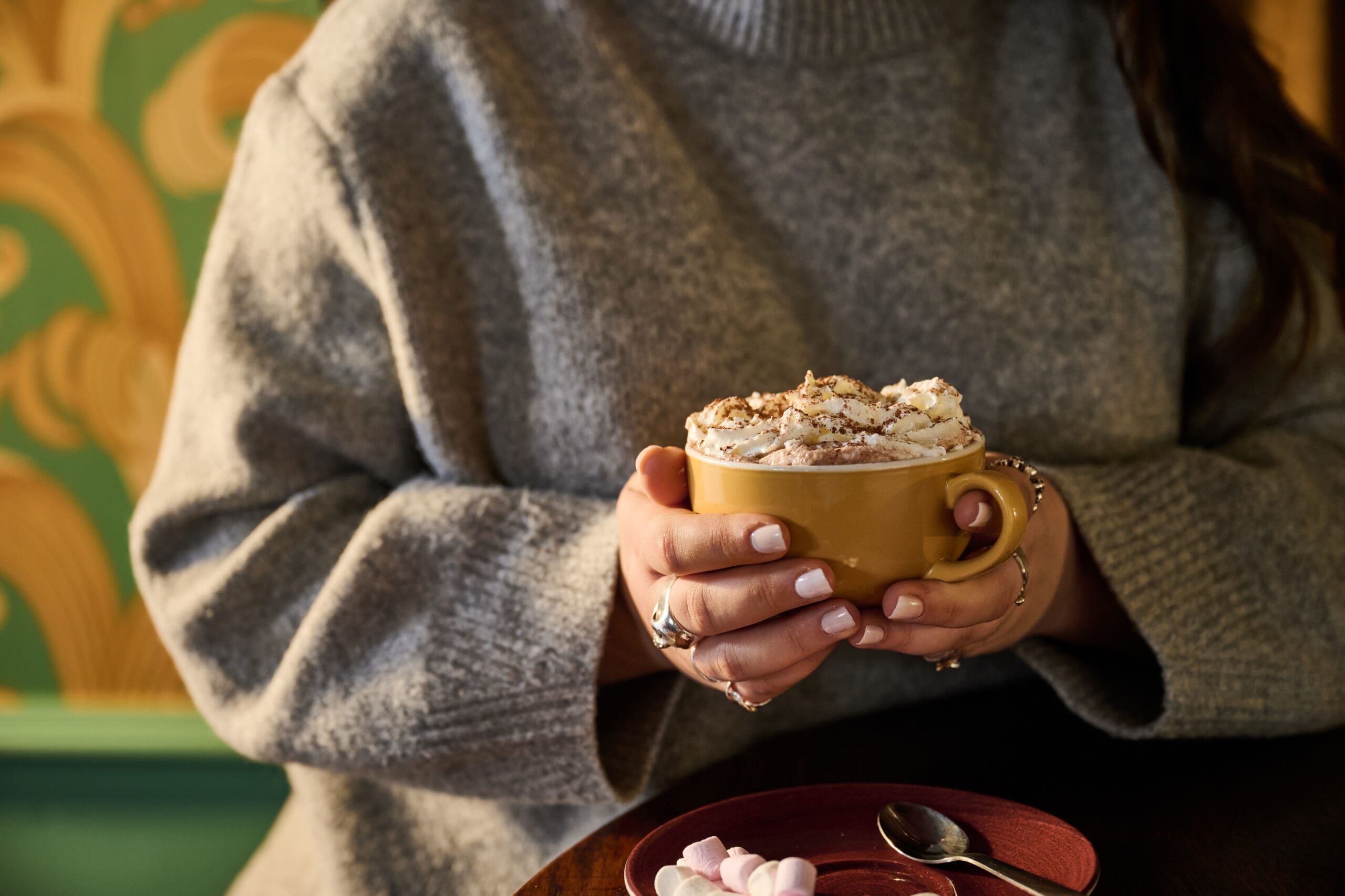 A person in a gray sweater lounges with a yellow mug topped with whipped cream. On the table, a red saucer holds small marshmallows and a spoon. The background features green and gold wallpaper.