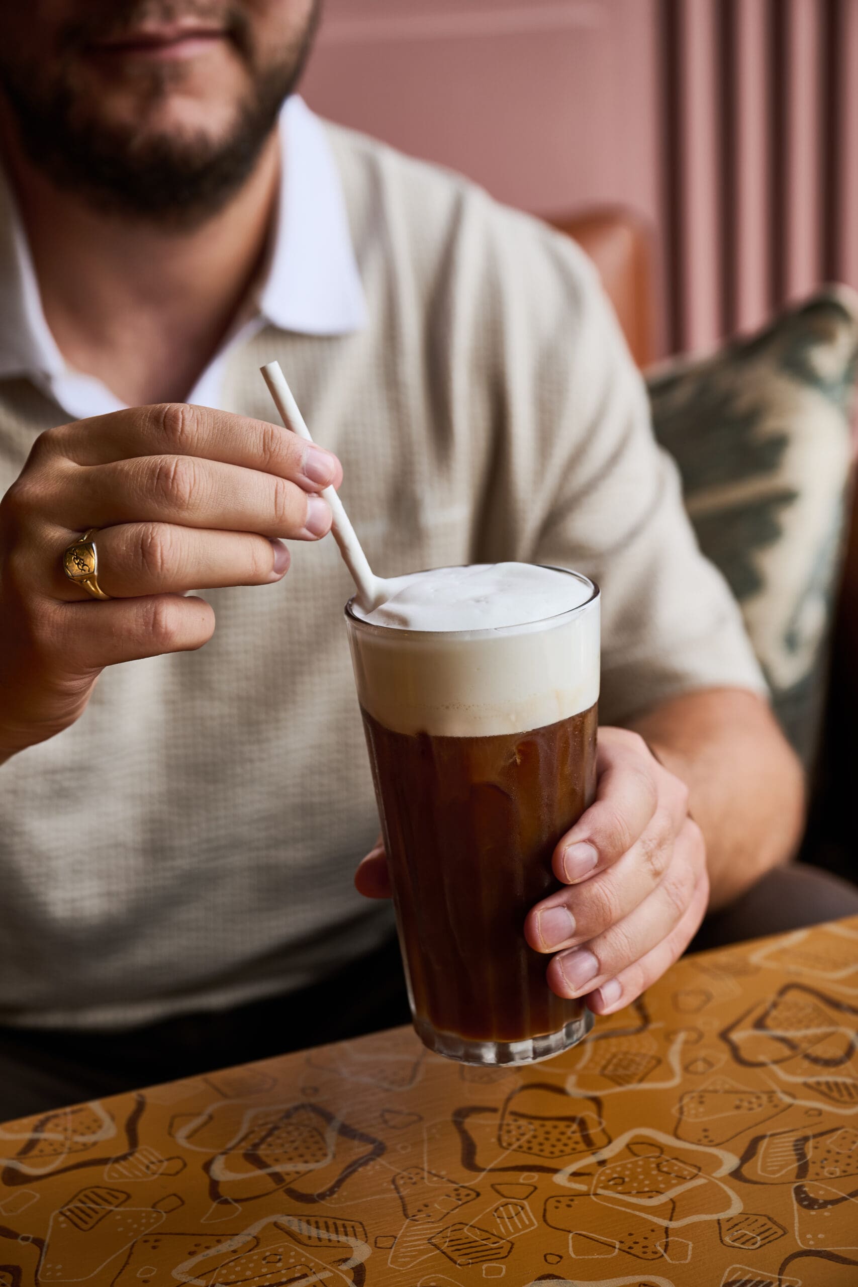 A person stirs a glass of iced coffee with a straw, topped with foam, while sitting at a patterned table. The person's face is partially visible, and they wear a beige shirt and a gold ring.