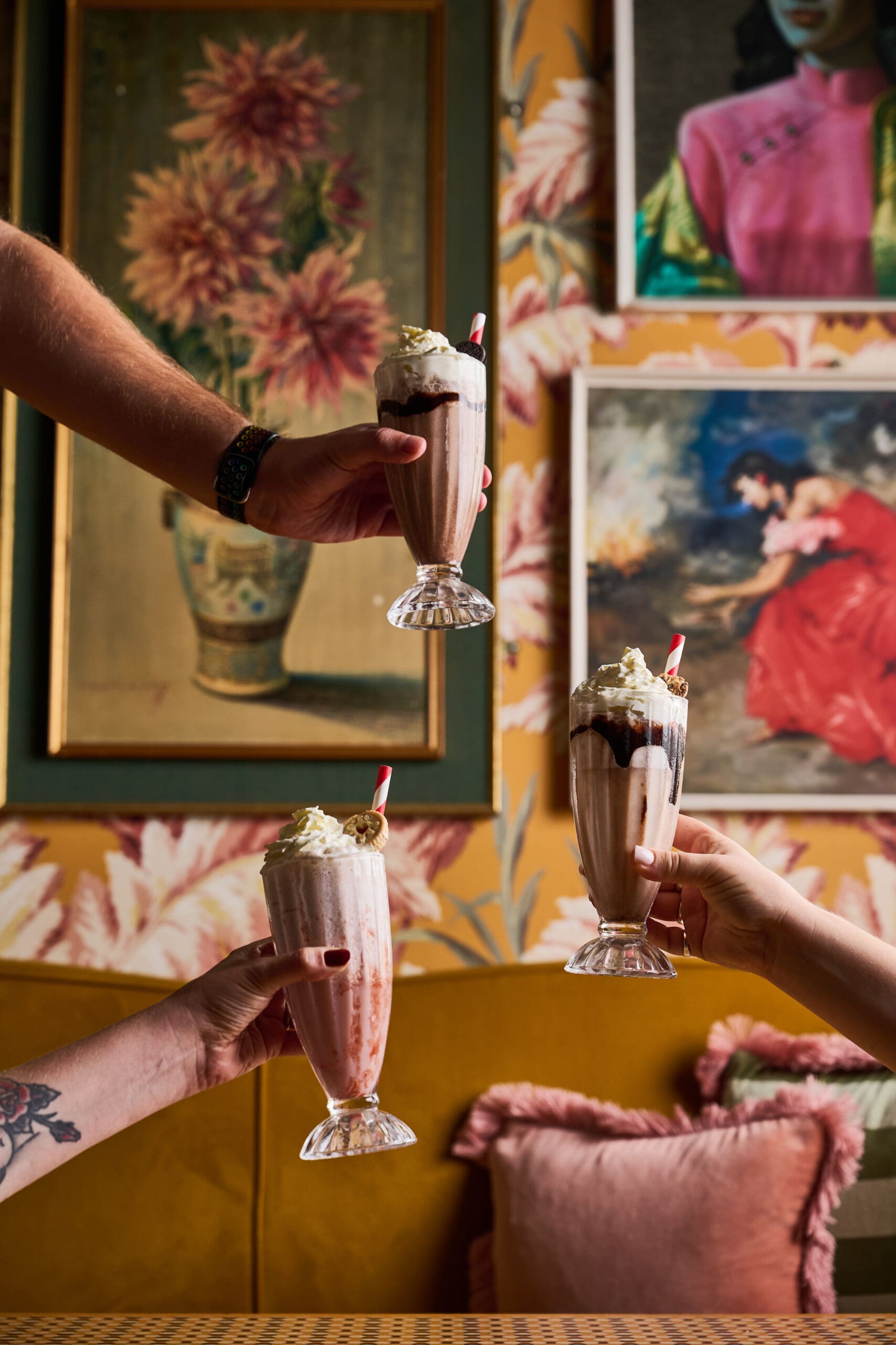 Three hands holding milkshakes with whipped cream and chocolate syrup, against a colorful, floral and art-filled wall with framed paintings and a yellow couch with pink pillows.