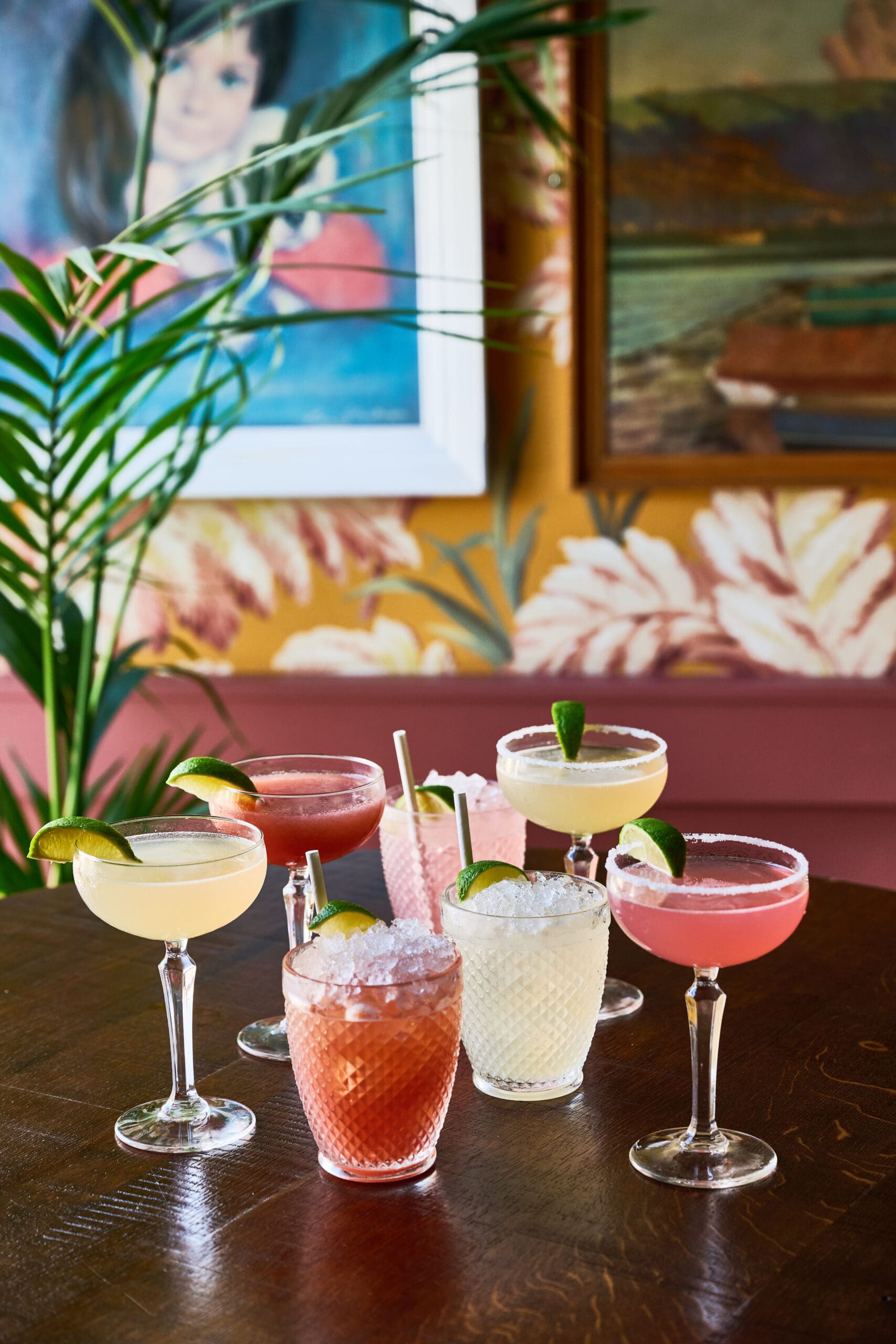 A group of colorful cocktails garnished with lime wedges and served in various glassware on a wooden table, with framed paintings and tropical wallpaper in the background.