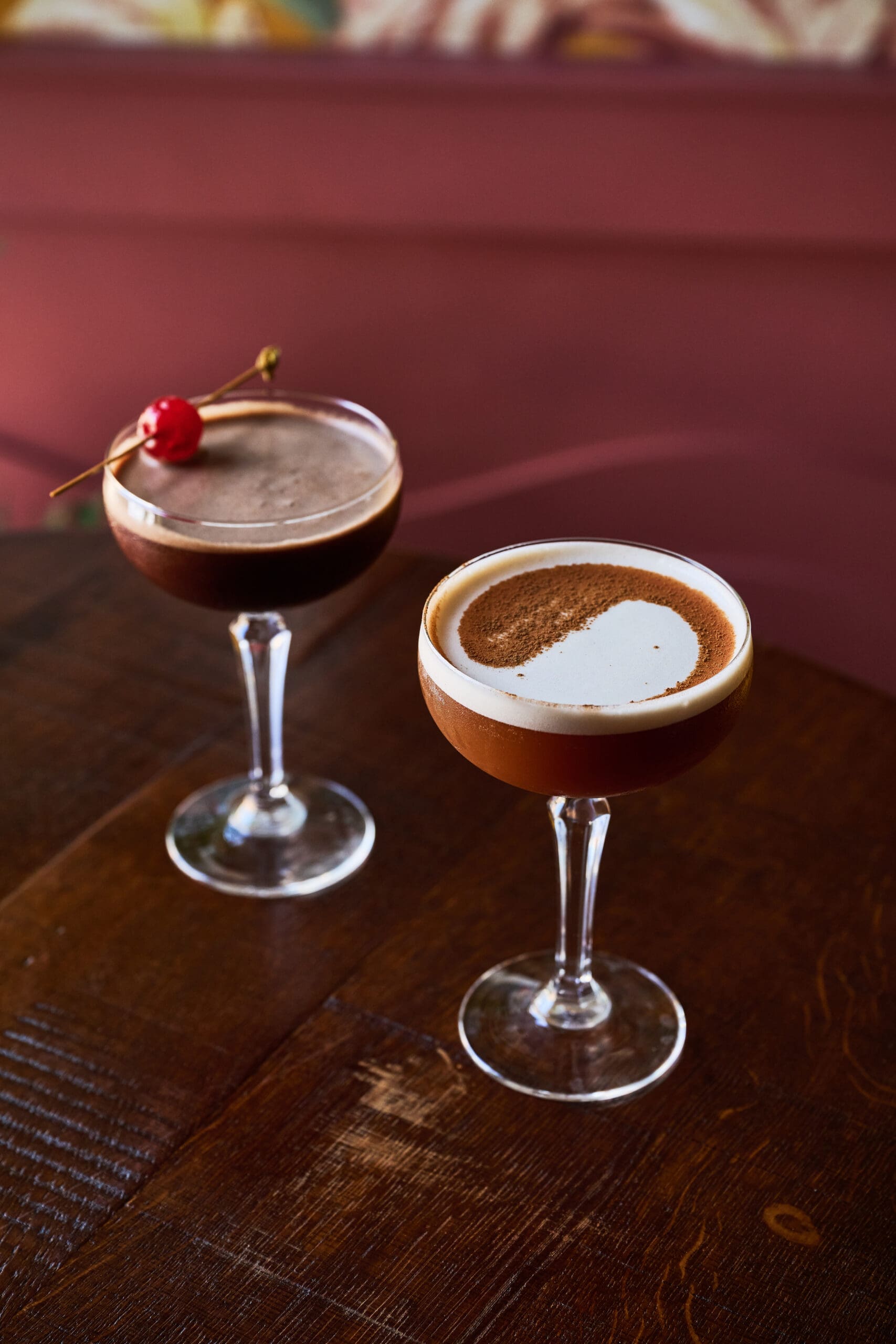 Two elegant coupe glasses on a wooden table, each filled with a dark cocktail. One is garnished with a cherry, and the other has a foamy top with a partial dusting of cocoa or coffee powder.