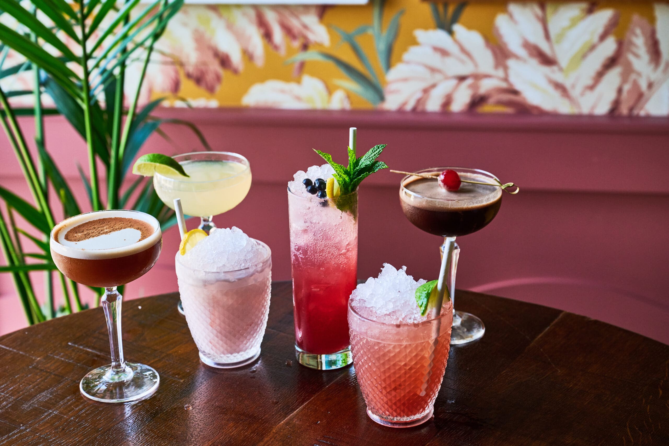 Five colorful cocktails in various glasses with fruit garnishes are arranged on a wooden table. The background features a leafy, tropical wallpaper and a green plant.