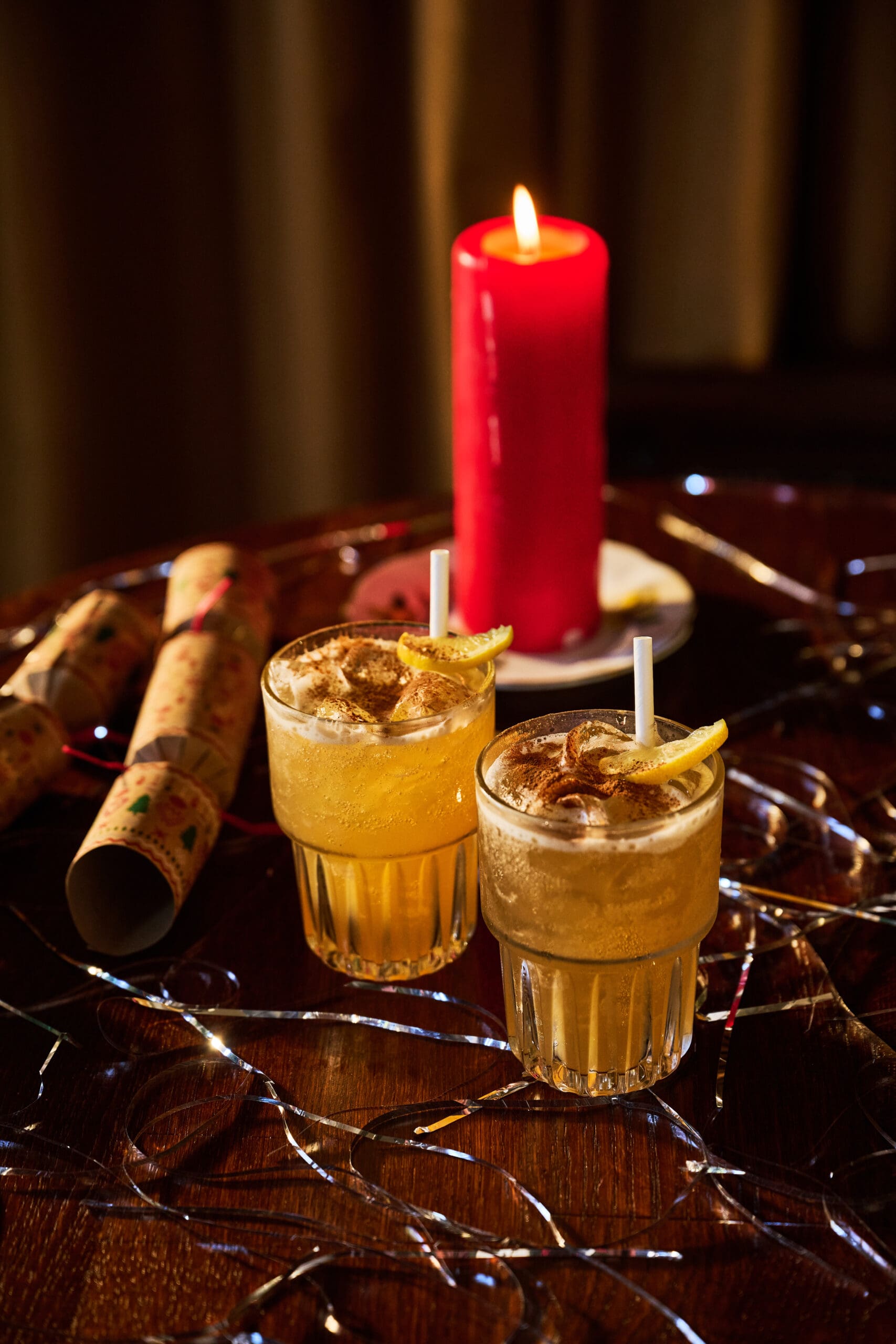 Two glasses of a yellow cocktail with lemon slices and straws sit on a table decorated with string lights, a Christmas cracker, and a lit red candle in the background.