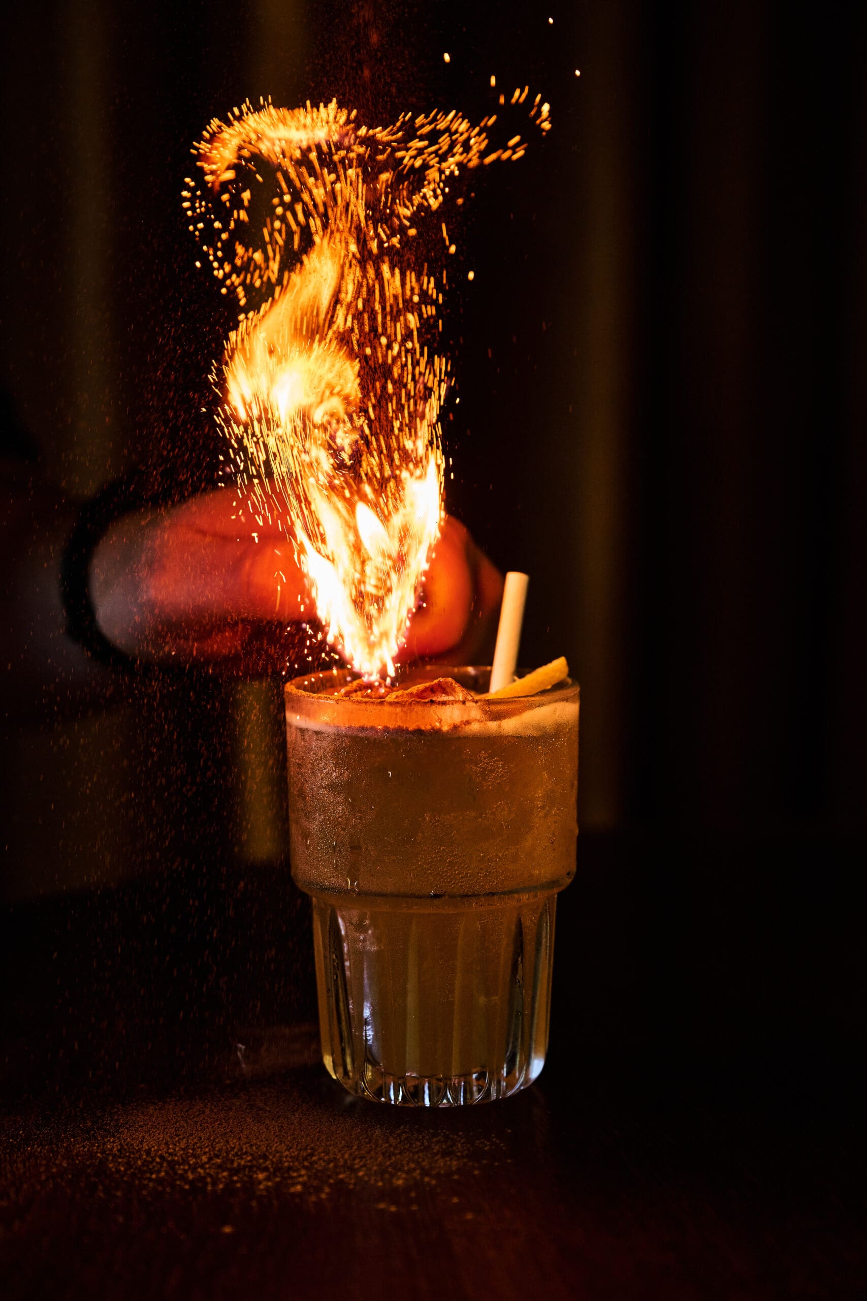 A cocktail in a glass emits bright sparks and flames, with a hand in the background igniting the drink. The dramatic fire effect stands out against a dark background, and a straw is visible in the glass.