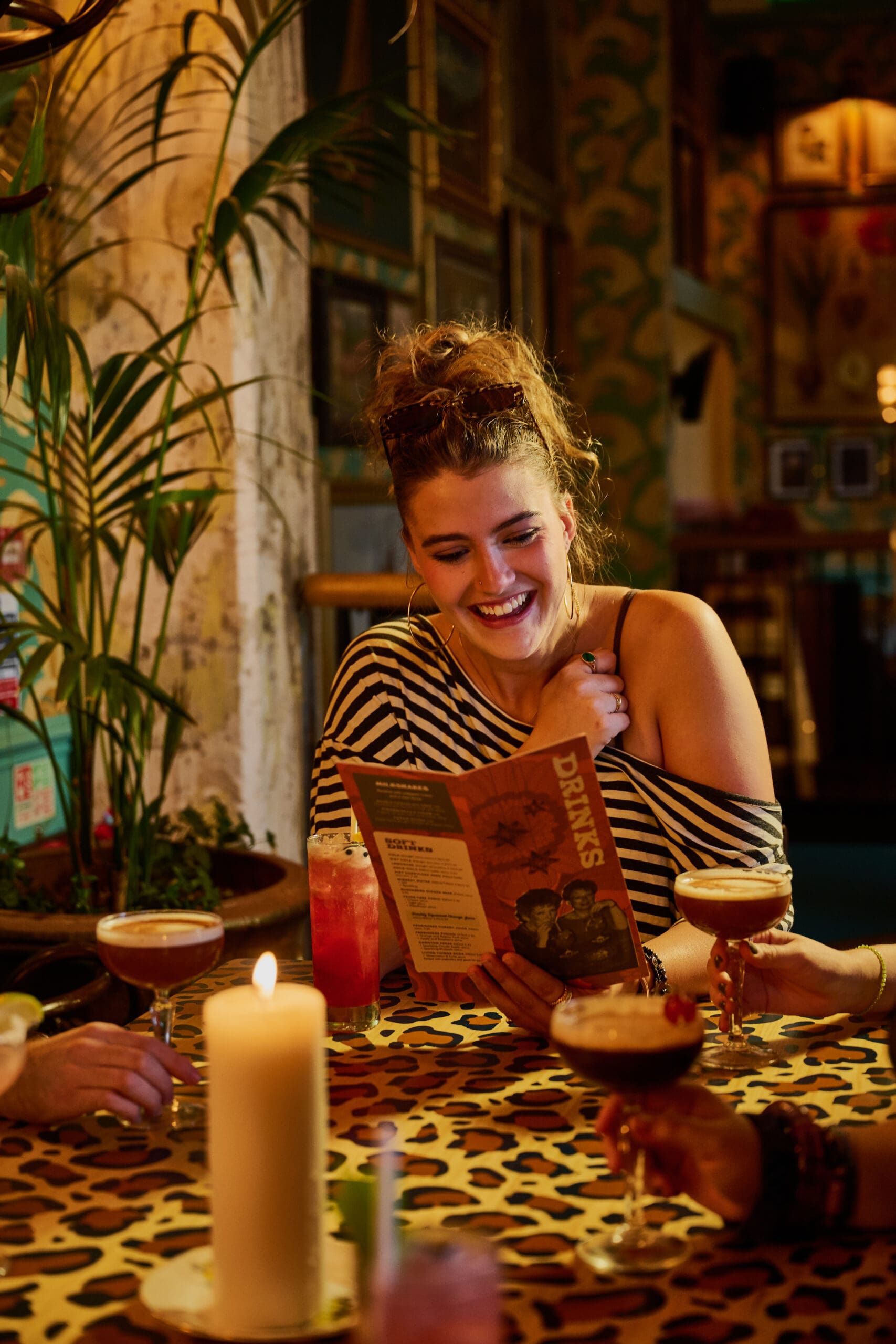 A smiling woman with curly hair sits at a leopard-print table in a dimly lit bar, holding a drinks menu. Cocktails and a lit candle are on the table. Other people’s hands reach in, holding drinks.