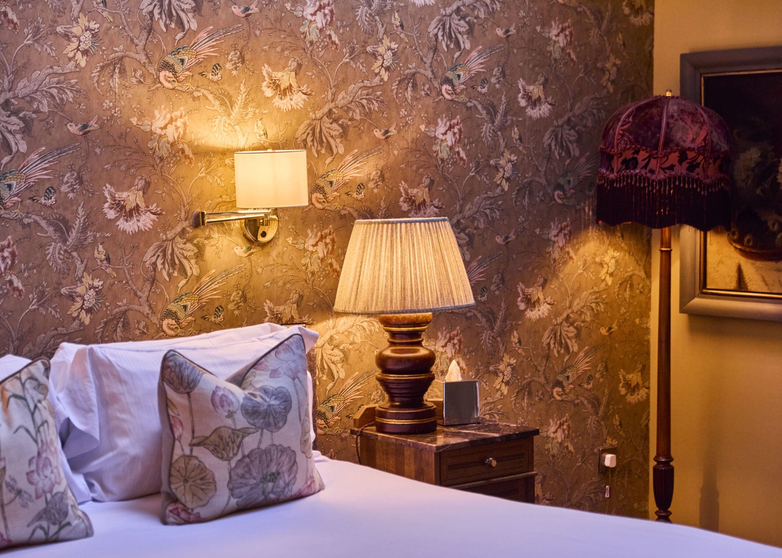 Vintage-style bedroom with floral wallpaper, a bed with patterned pillows, a wooden nightstand with a lamp, a wall sconce, a velvet floor lamp, and part of a framed painting. Warm, cozy lighting fills the room.