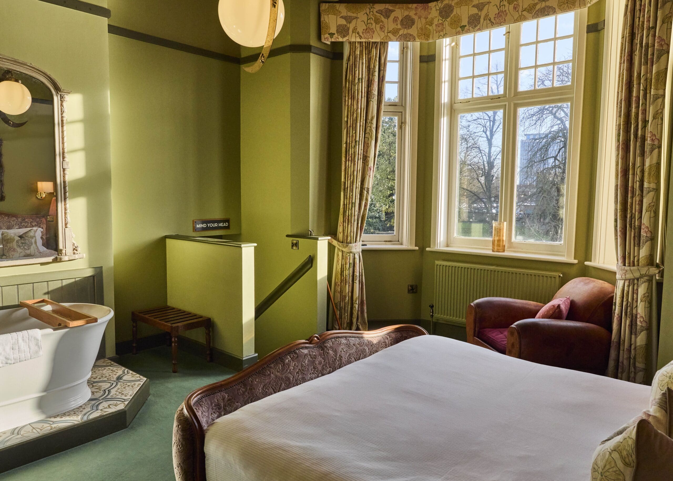 A sunlit bedroom with pale green walls, a large window with curtains, an armchair, a bed with a wooden frame, a small wooden bench, and a freestanding bathtub near a decorative mirror.