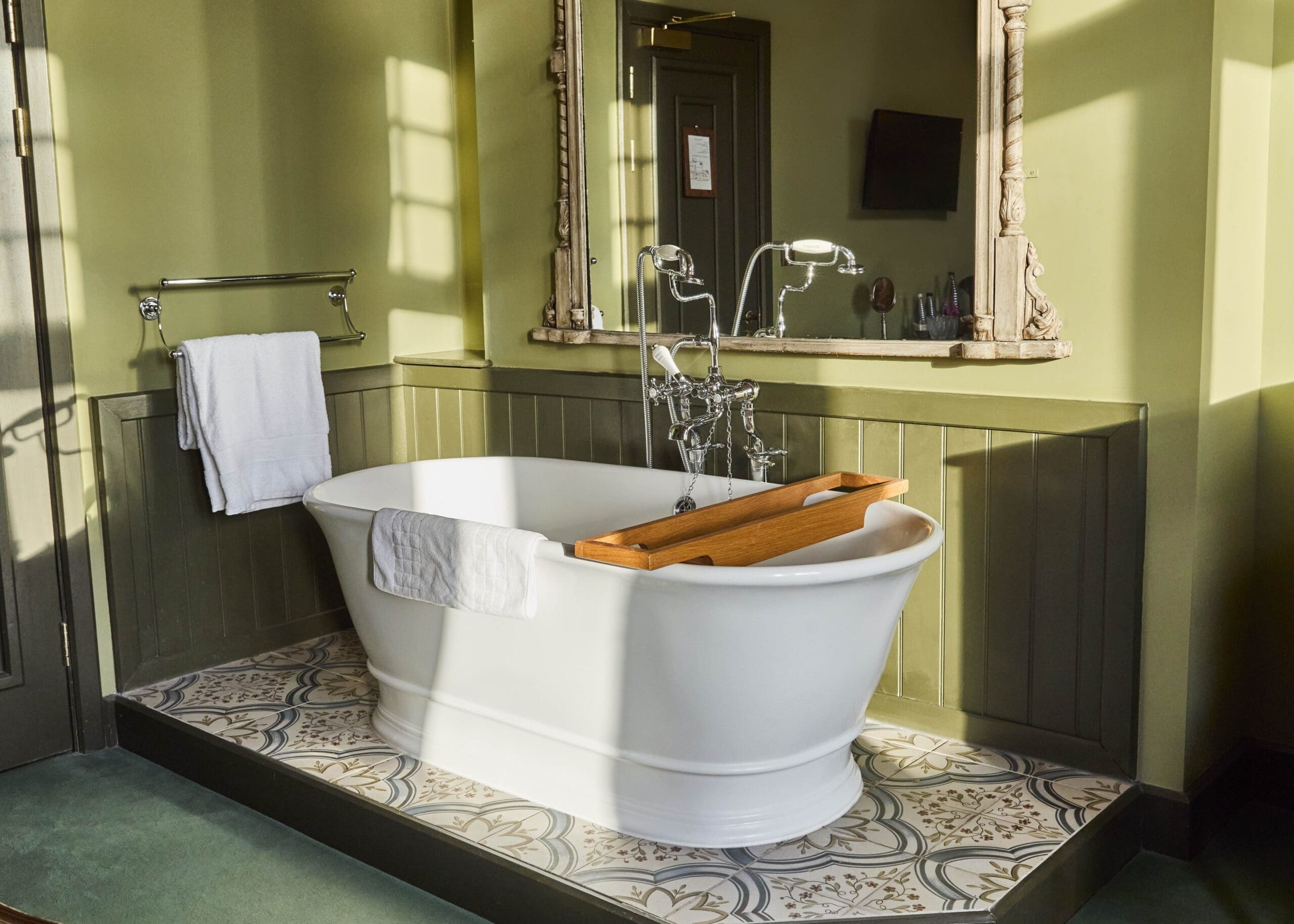 A white freestanding bathtub with a wooden bath tray and towel sits on decorative tile flooring in a sunlit bathroom with green walls, a large mirror, and a towel rack.