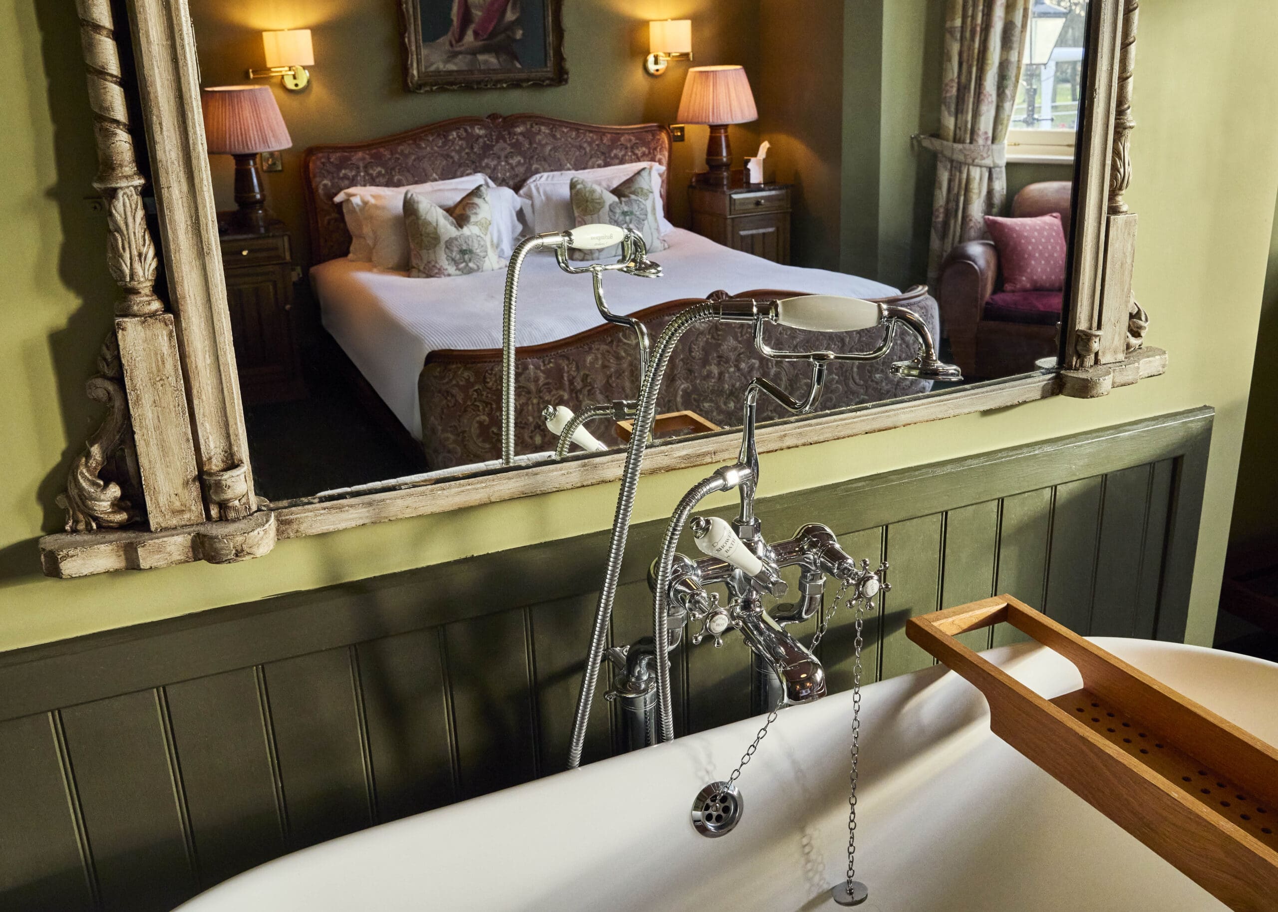 A vintage-style bathtub with a wooden tray sits in front of a large mirror reflecting a cozy bedroom with a double bed, lamps, and pillows, creating a warm, inviting atmosphere.