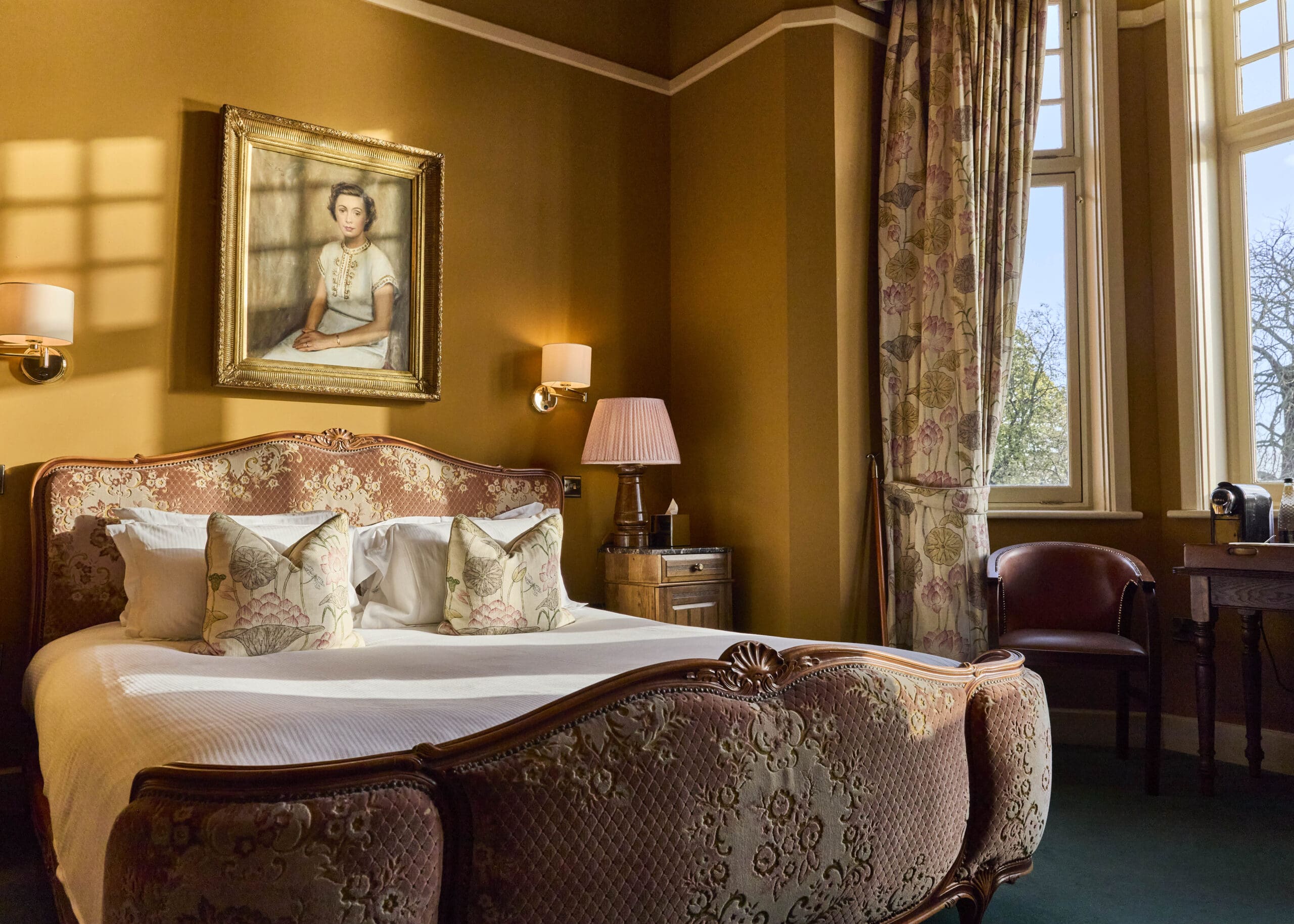 A warmly lit vintage bedroom with a patterned bed, floral pillows, a side table with a lamp, large windows with curtains, a desk, and a framed portrait of a young woman hanging above the bed.