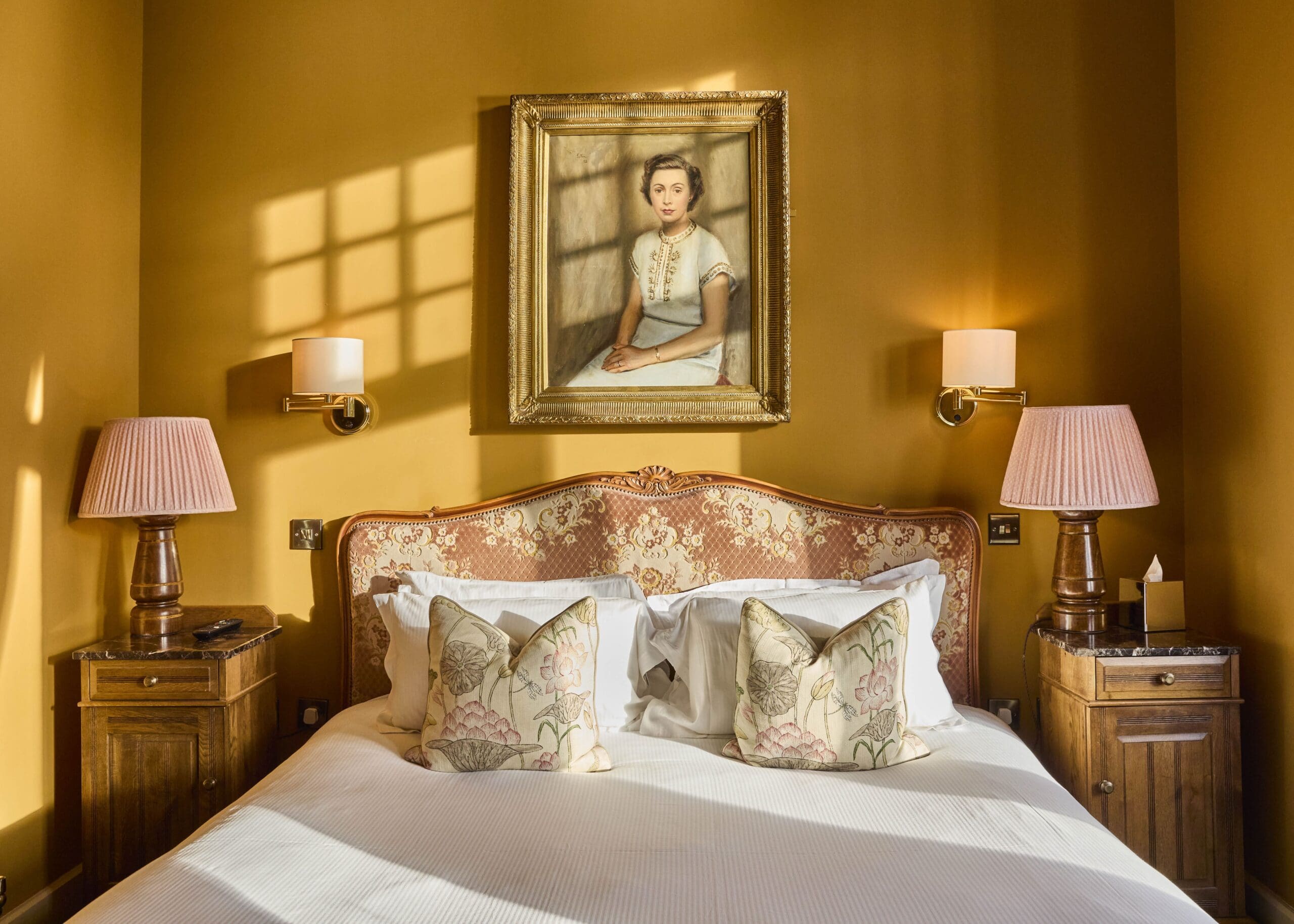 A cozy bedroom with golden-yellow walls, a framed portrait of a woman above a vintage upholstered bed, two floral pillows, matching wooden nightstands, and pink lamps on each side. Sunlight casts window shadows on the wall.