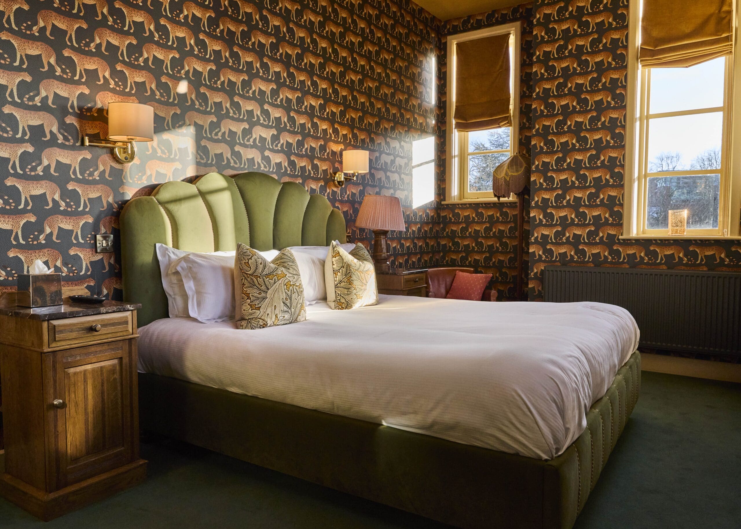 A stylish bedroom with a large green upholstered bed, white bedding, decorative pillows, wooden bedside table, and walls covered in cheetah-patterned wallpaper. Sunlight streams in through two windows with matching blinds.