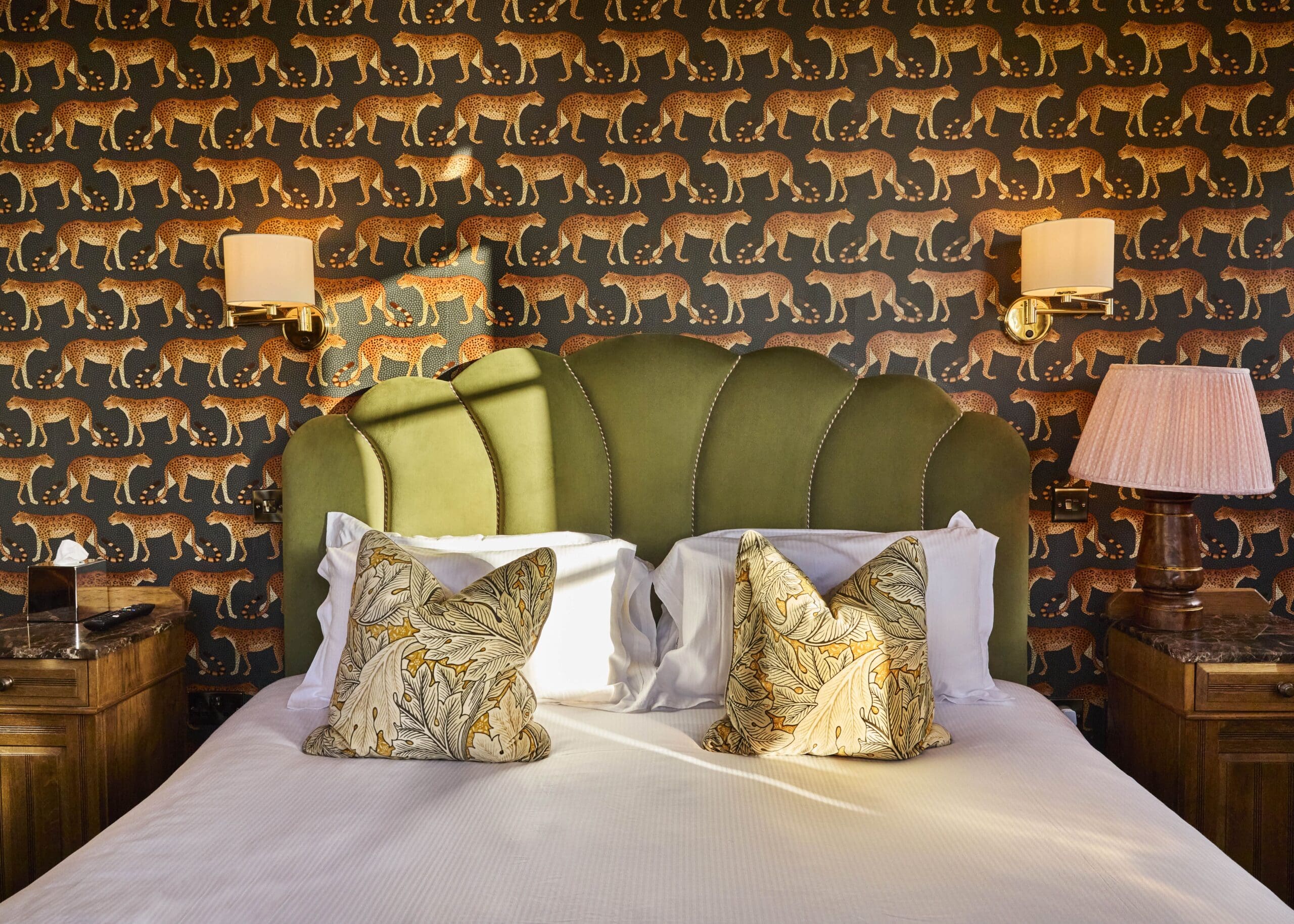 A neatly made bed with a green, scalloped headboard sits against a wall with a repeating cheetah wallpaper pattern. Two patterned pillows are on the bed, with wall lamps and wooden nightstands on either side.