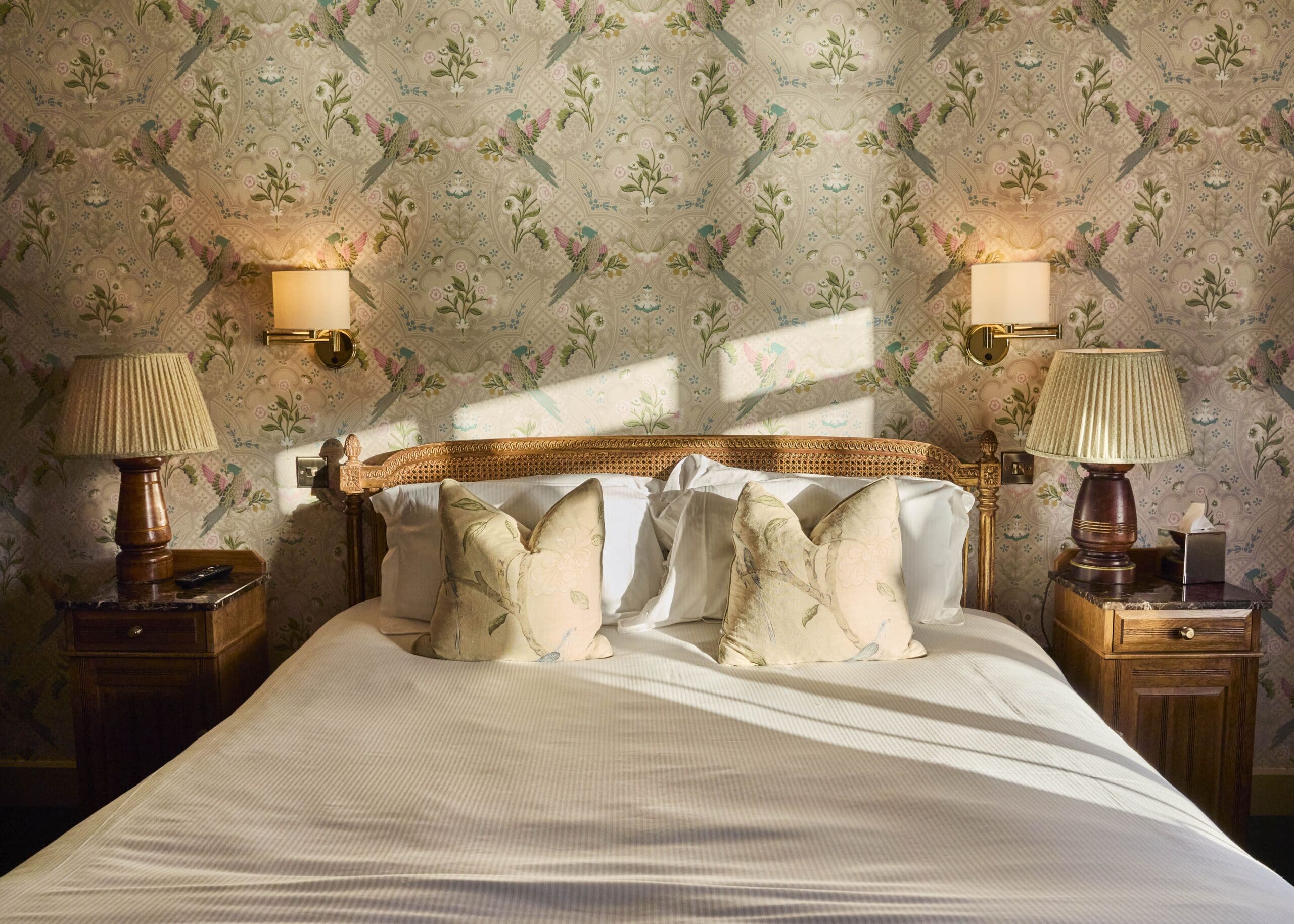 A neatly made bed with light-colored pillows and a white duvet sits between two wooden nightstands, each with a lamp. Floral-patterned wallpaper and two wall-mounted lights decorate the background. Sunlight casts shadows on the bed.