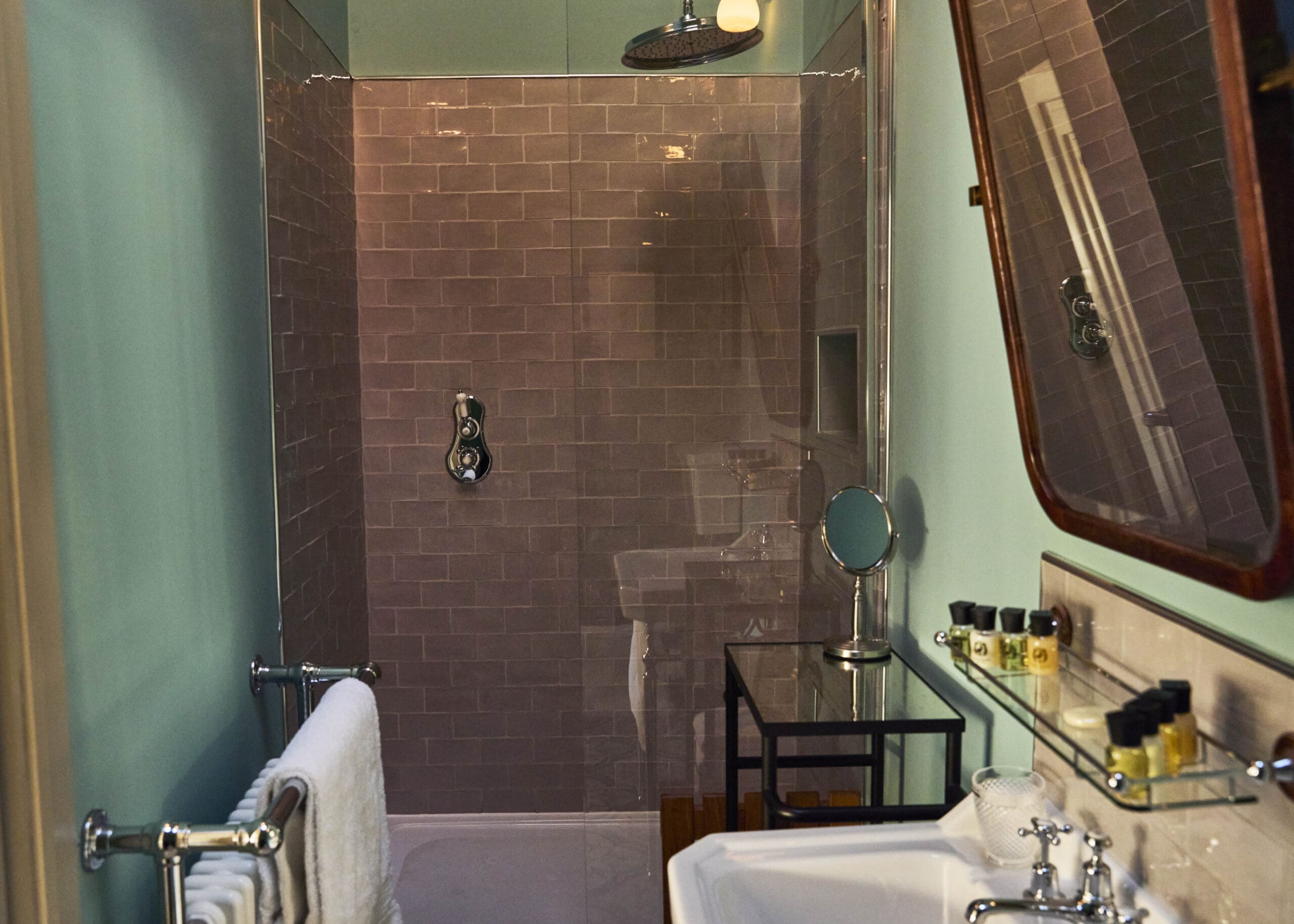 A modern bathroom with a glass shower enclosure, gray subway tiles, a chrome rain showerhead, a towel rack with folded towels, a black metal shelf, and a sink with toiletries on a glass shelf.