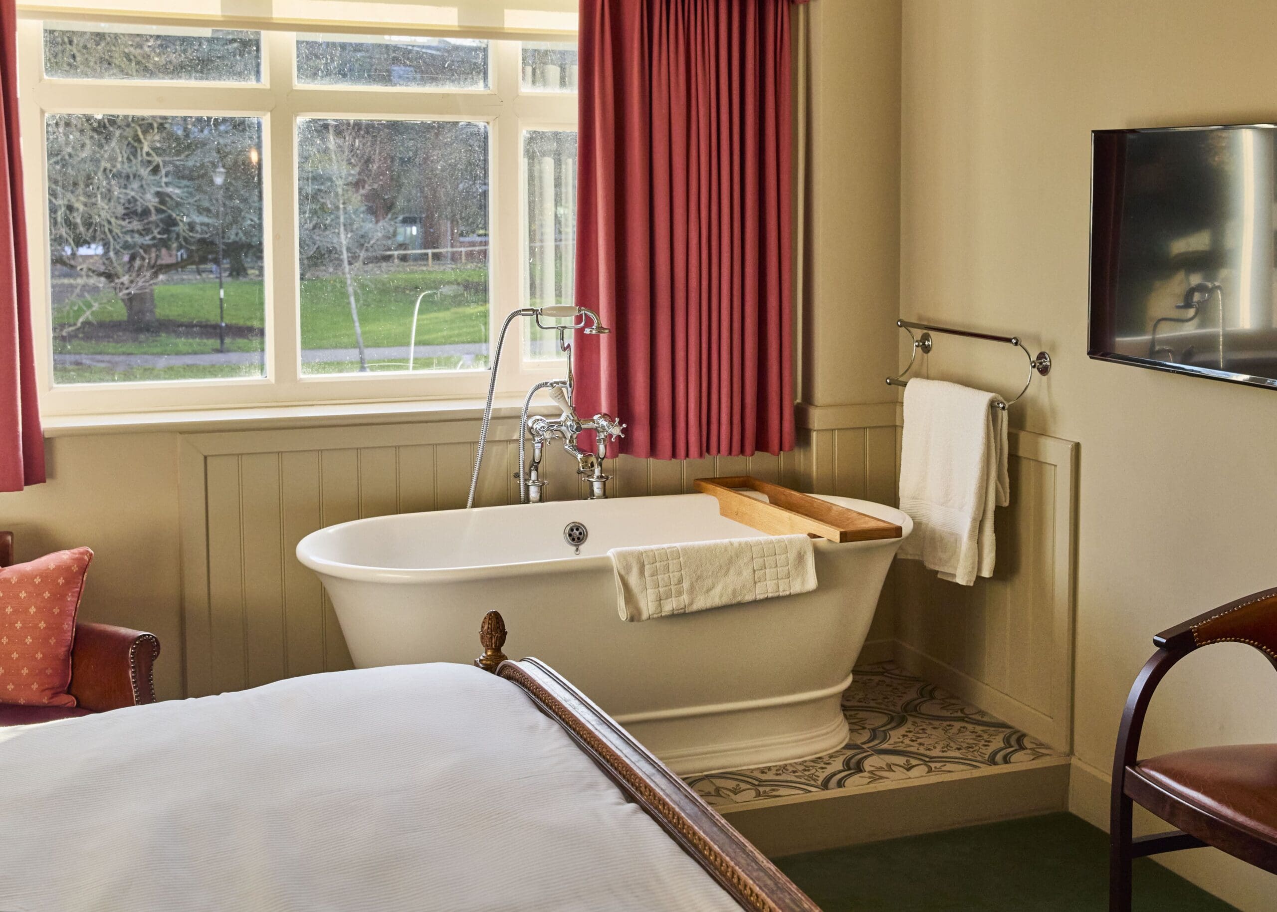 A cozy bathroom with a freestanding white bathtub beside a window, red curtains, a towel rack with white towels, and a wooden bath tray. The scene includes part of a bed and a view of trees outside.