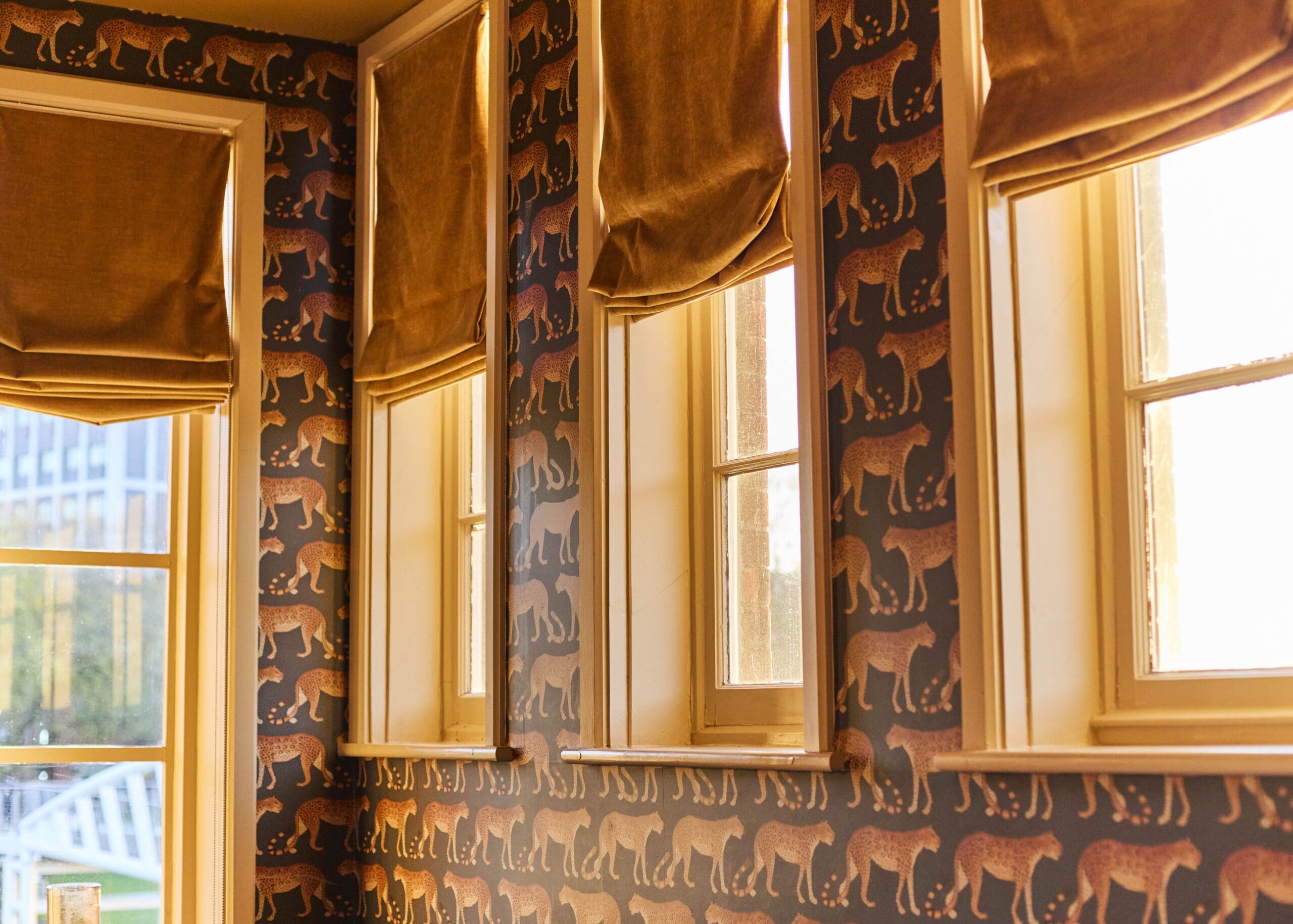 Three sunny windows with tan roman shades line a wall decorated with wallpaper featuring a pattern of leopards walking in rows. Warm daylight streams through, creating a cozy atmosphere.