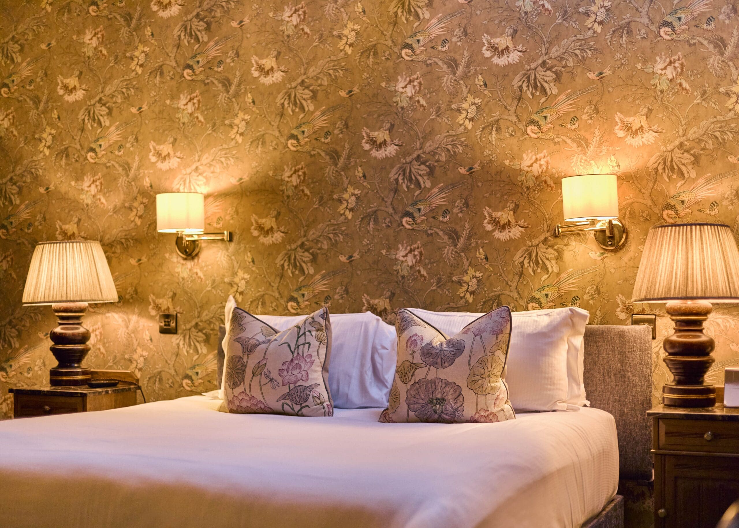 A neatly made bed with two floral pillows, flanked by two bedside tables with lamps, and two wall-mounted lights. The background features ornate, floral-patterned wallpaper in warm tones.