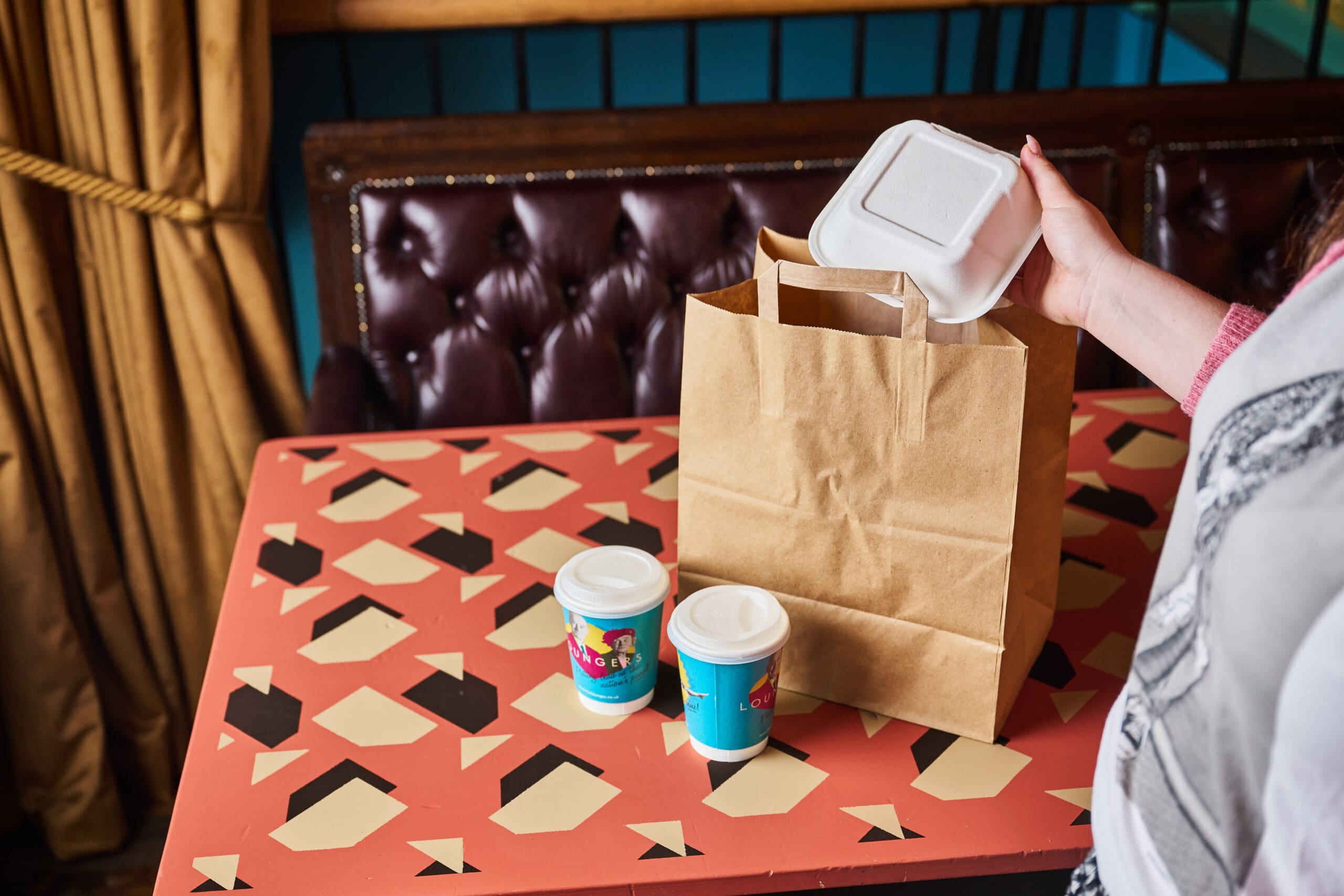 A person places a white takeout container into a brown paper bag on a patterned table with two takeaway coffee cups nearby.