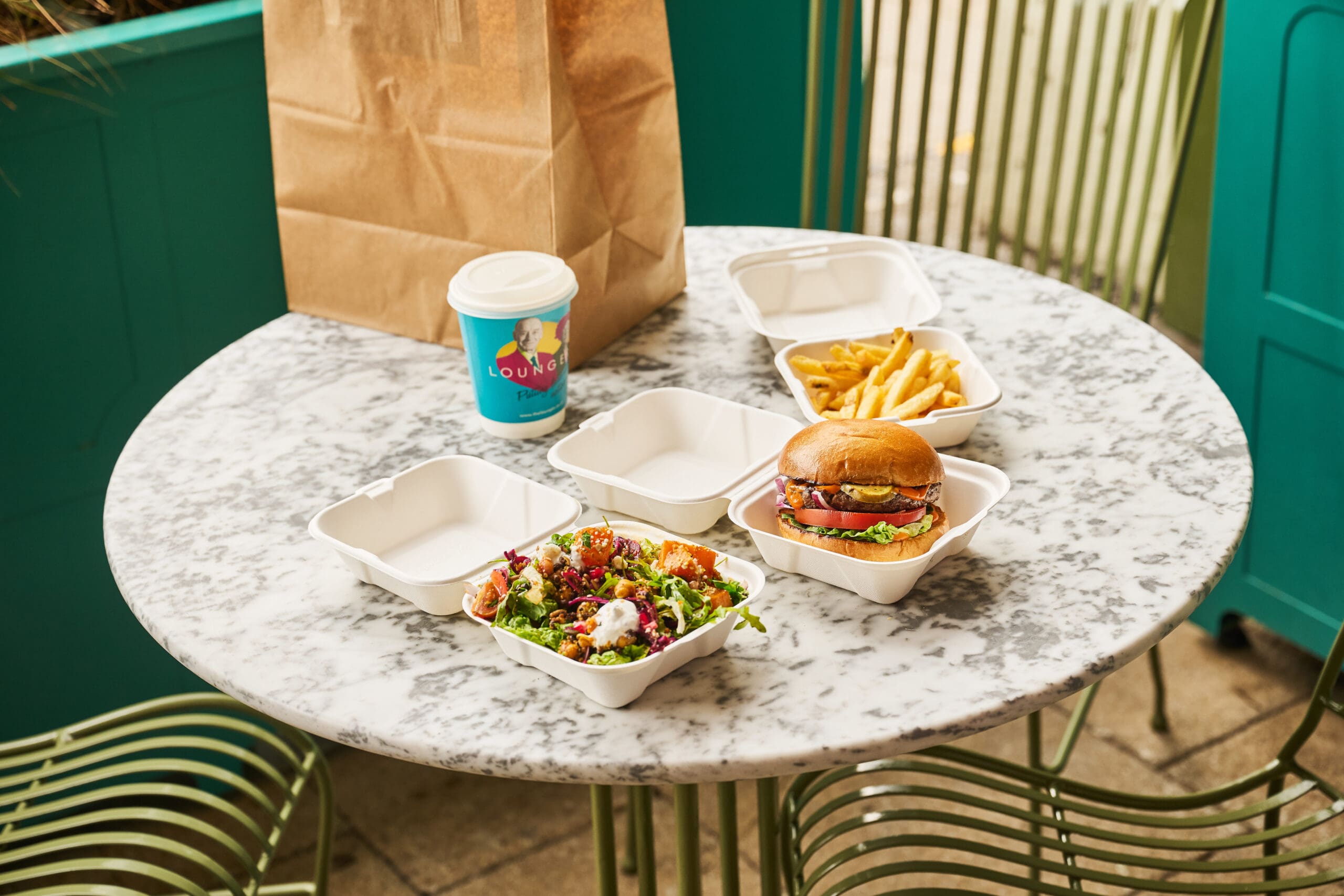 A marble table holds a takeout burger with lettuce and tomato, fries, a fresh salad with colorful vegetables, a paper coffee cup, a brown paper bag, and empty takeout boxes, set outside by green chairs and a teal wall.