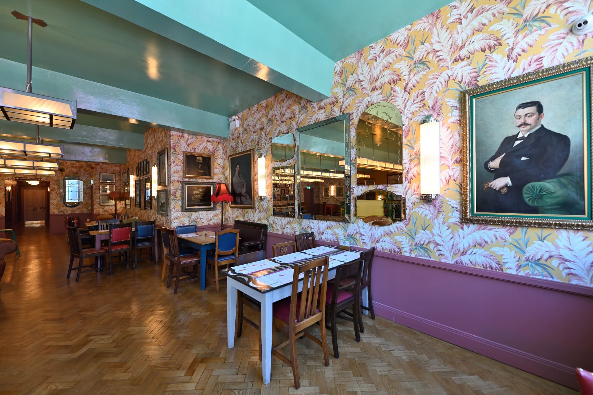 A vintage-style restaurant interior at Lupo features colorful wallpaper, mismatched tables and chairs, large mirrors, framed paintings—including a mustached portrait—and wooden parquet flooring.