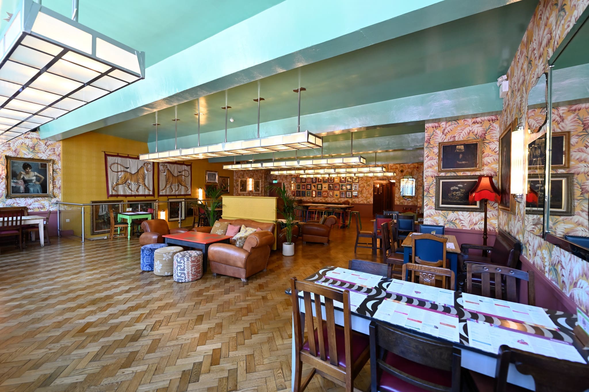 A stylish, eclectic restaurant interior—Lupo—features patterned wallpaper, wooden flooring, leather sofas, colorful chairs, contemporary lighting, and framed art. Tables are set and the vibrant space feels inviting.