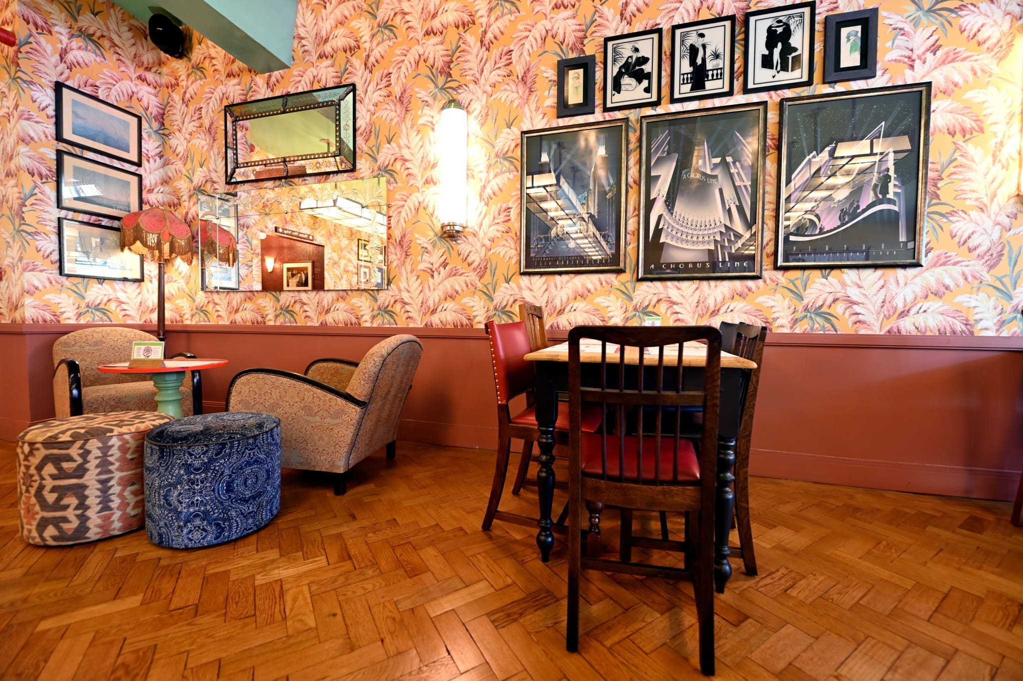 A cozy, retro-styled Lupo cafe interior with patterned wallpaper, eclectic framed art, vintage armchairs, a wooden table with chairs, round ottomans, and a decorative lamp on a parquet floor.