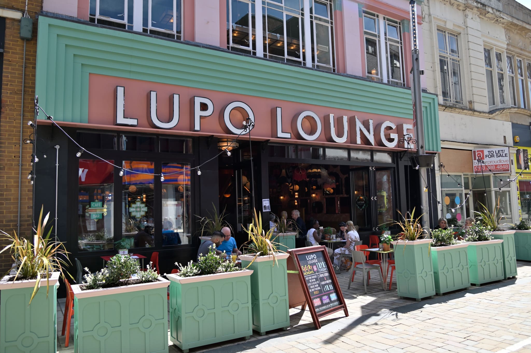 Outdoor view of Lupo Lounge café with large Lupo sign, pastel green and pink facade, people sitting at tables outside, and planter boxes with flowers lining the front on a sunny day.