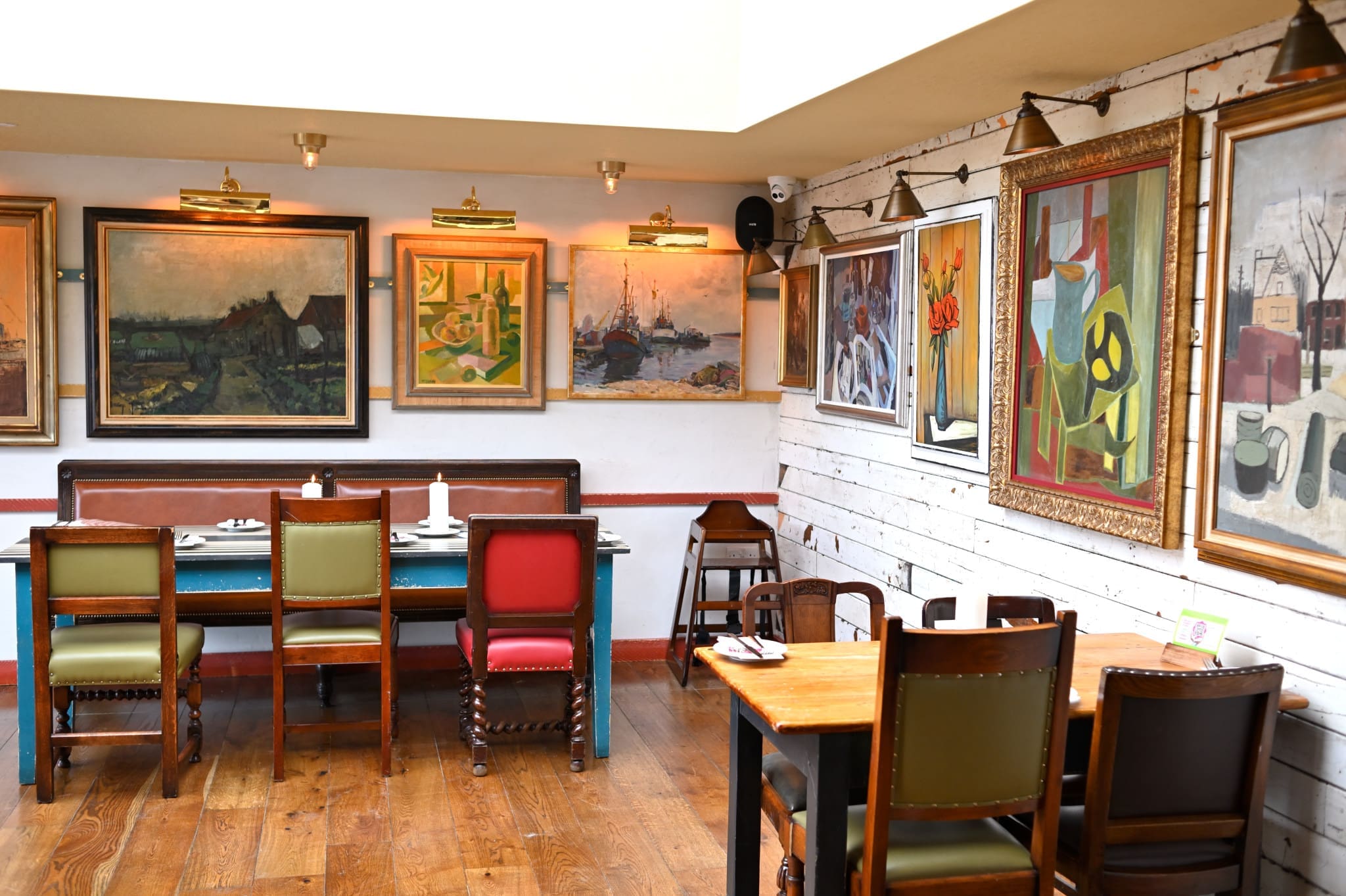 A cozy Marisco restaurant interior with wooden floors, colorful chairs, tables set for dining, and walls decorated with various large paintings in different styles and frames. Warm lighting illuminates the artwork.