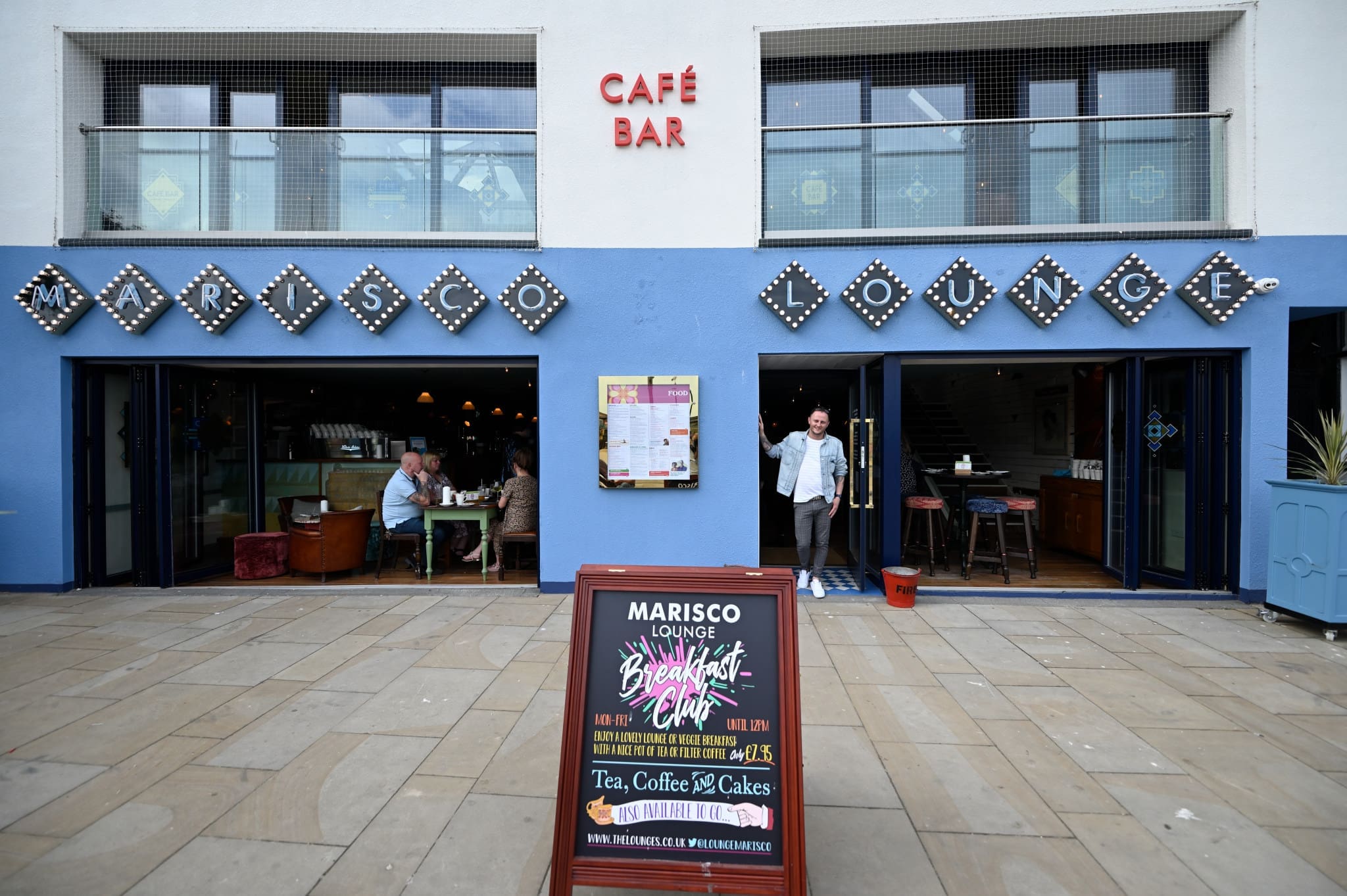 The exterior of Marisco Lounge café bar, with open doors revealing people dining inside; a man stands outside, and a signboard advertises Marisco’s breakfast, tea, coffee, and cakes.