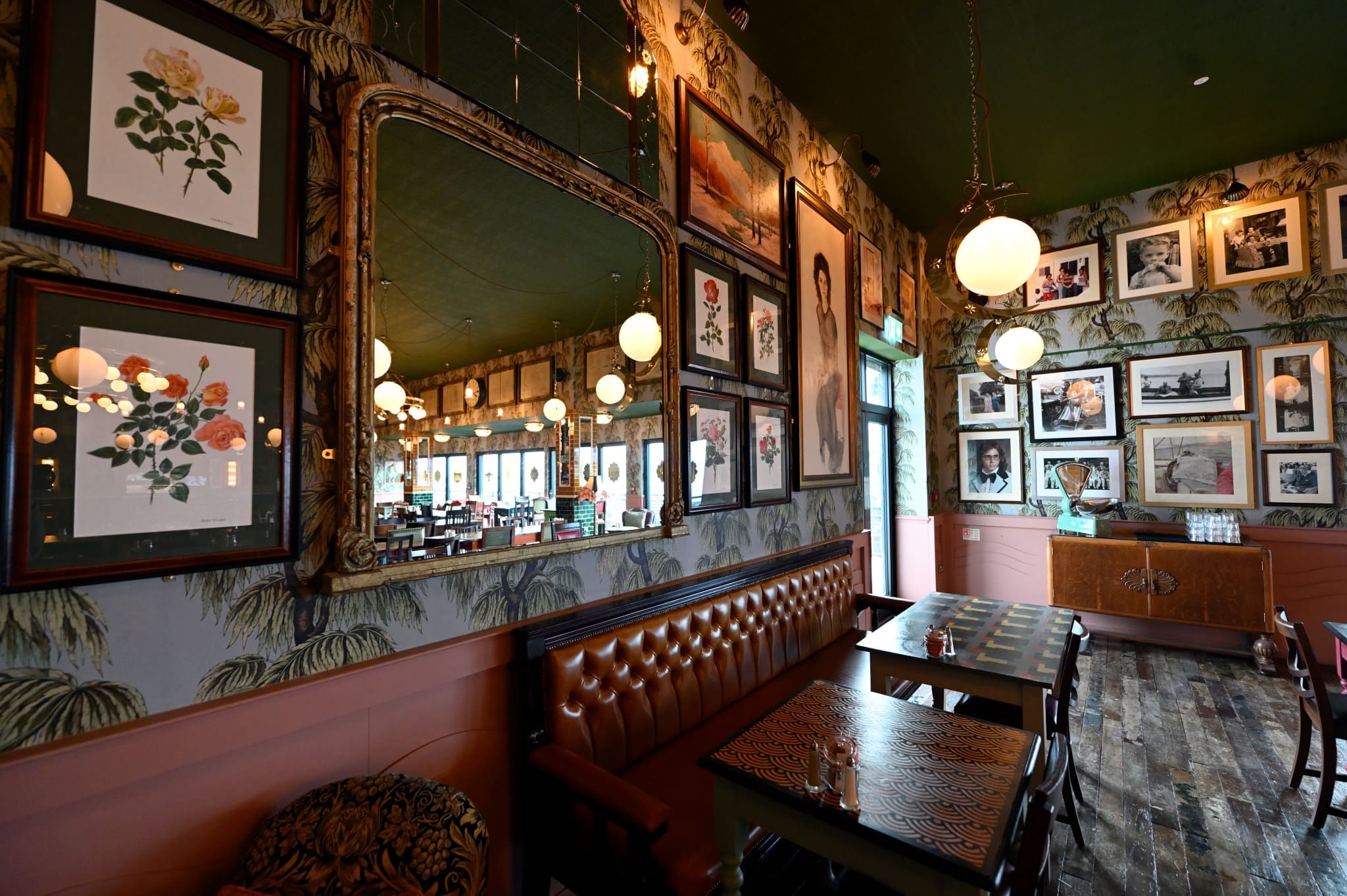 A stylish Martello restaurant interior with patterned wallpaper, framed art and photographs on the walls, leather bench seating, wooden tables, globe pendant lights, and a large ornate mirror reflecting the room.
