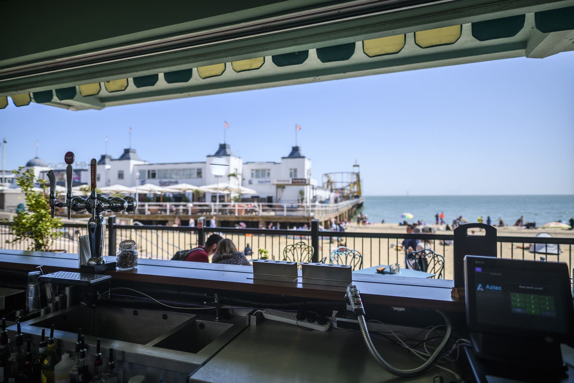 View from behind a bar looking out to a sunny beach scene with people, white buildings, and the iconic Martello tower near the pier by the sea under a clear blue sky. The bar counter and equipment are visible in the foreground.
